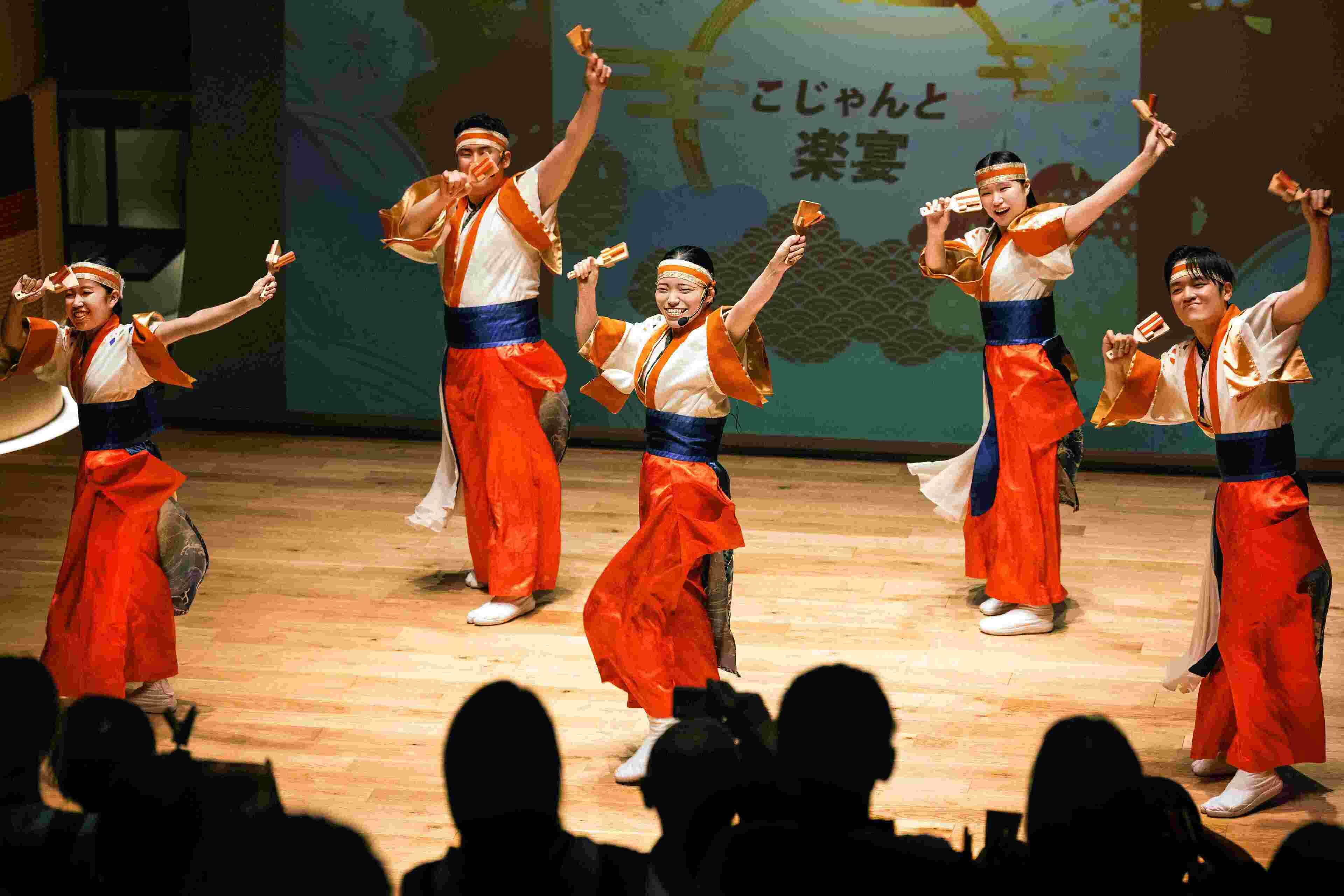 A group of performers in traditional costumes on a stage with a wooden floor and a backdrop with Japanese text.
