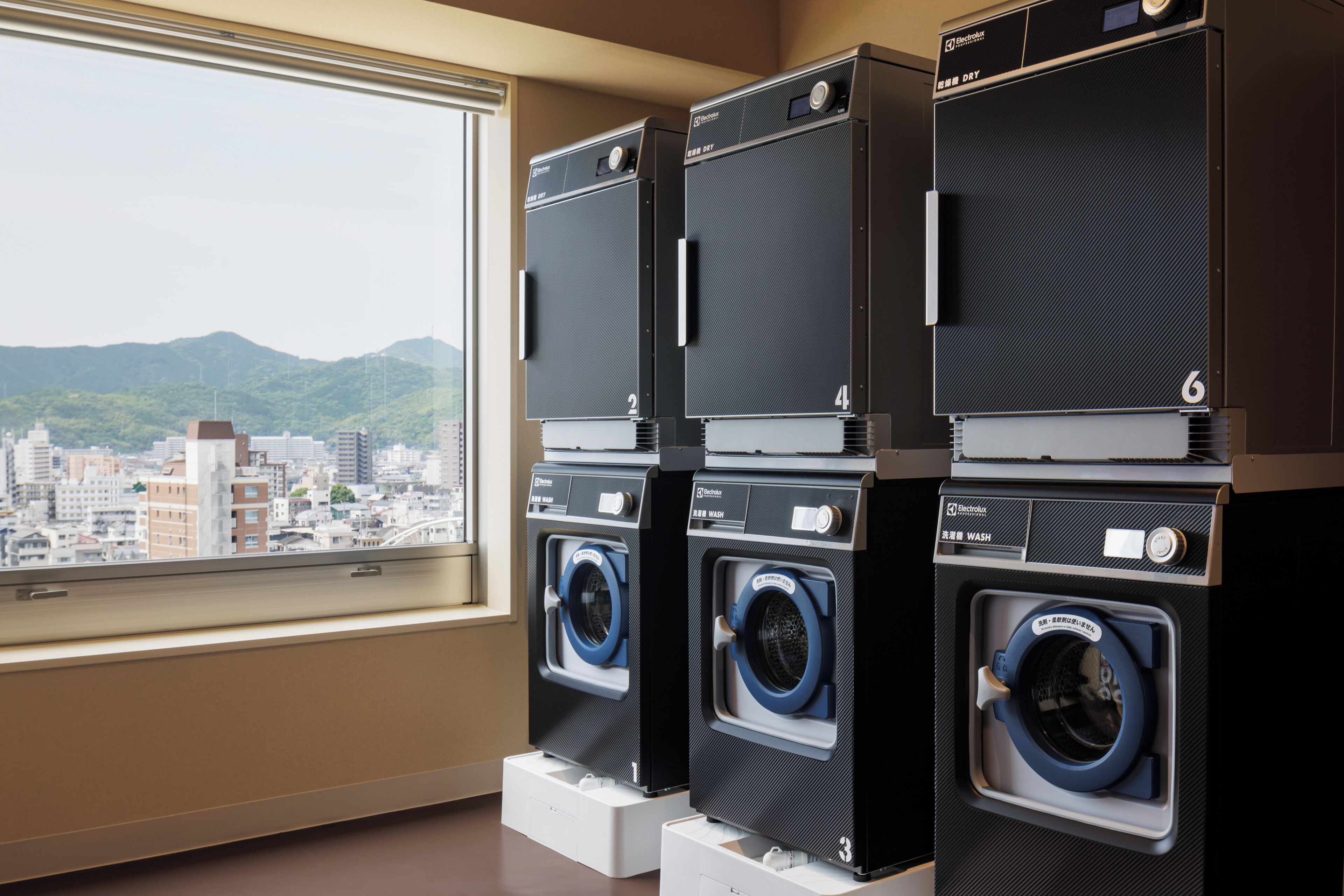 A row of stacked laundry machines in a room with a large window overlooking a cityscape.