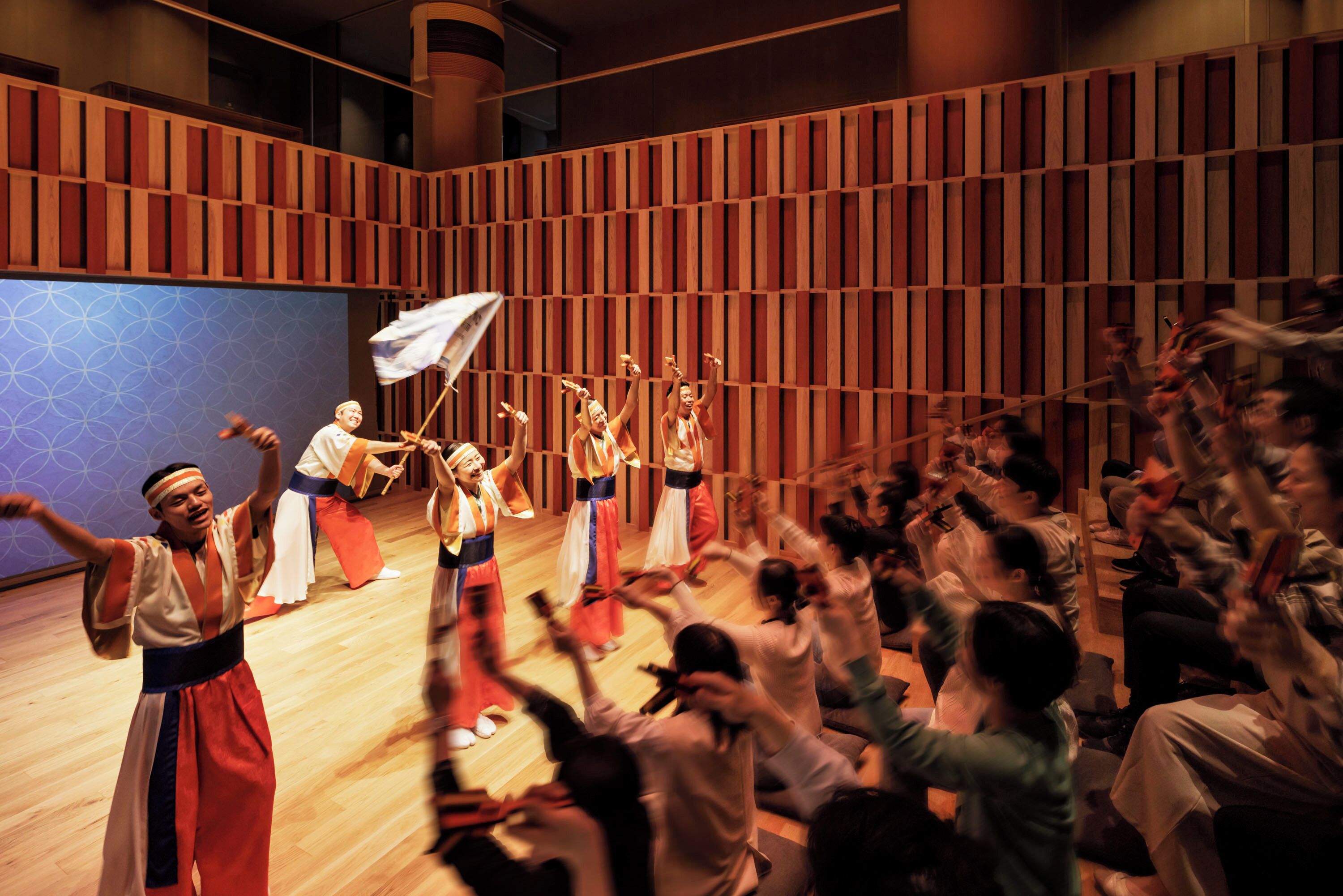 A group of people in traditional attire performing on a stage with a wooden backdrop and a patterned wall.