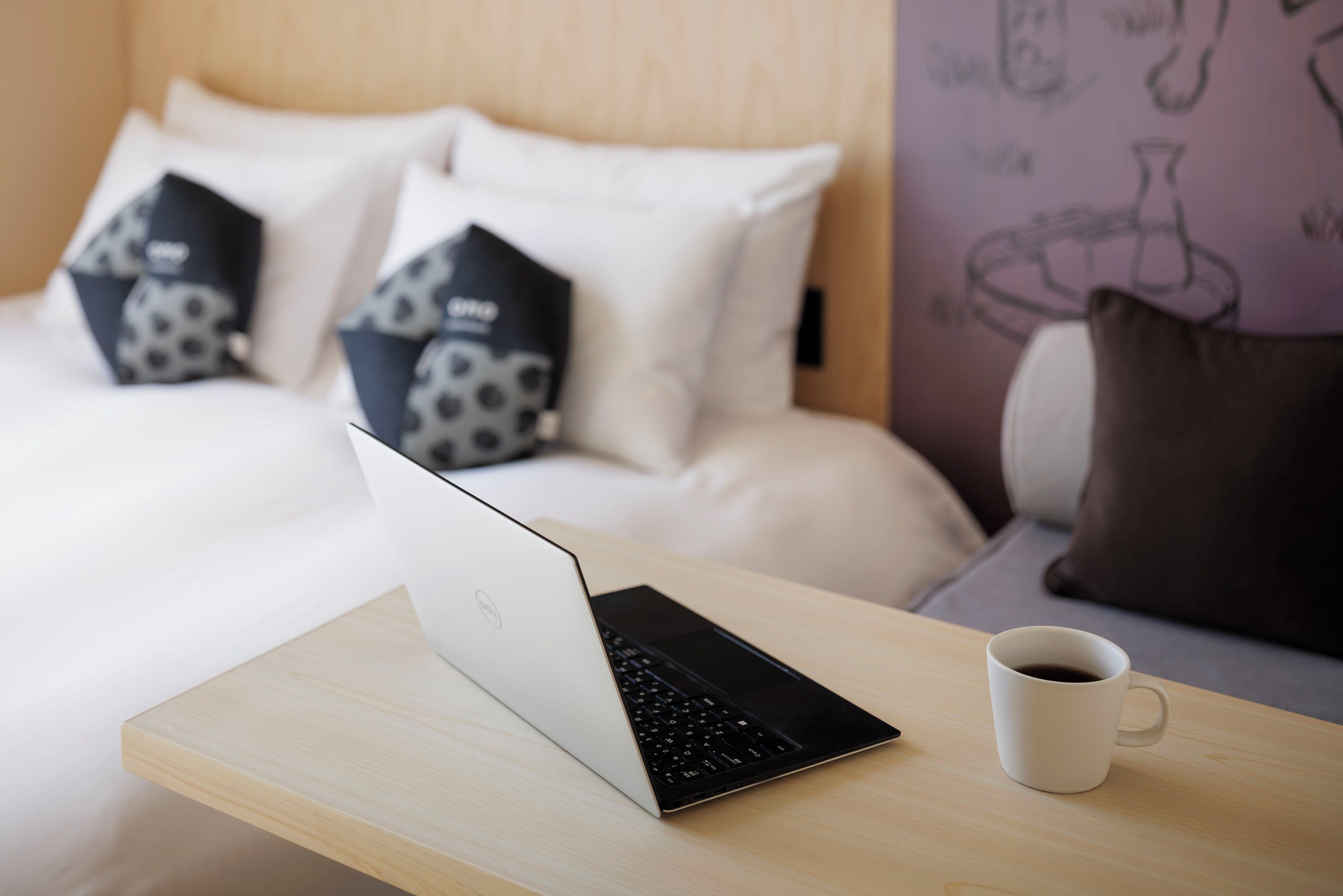 A laptop on a wooden table with a white mug beside it, in a room with a bed and patterned pillows.