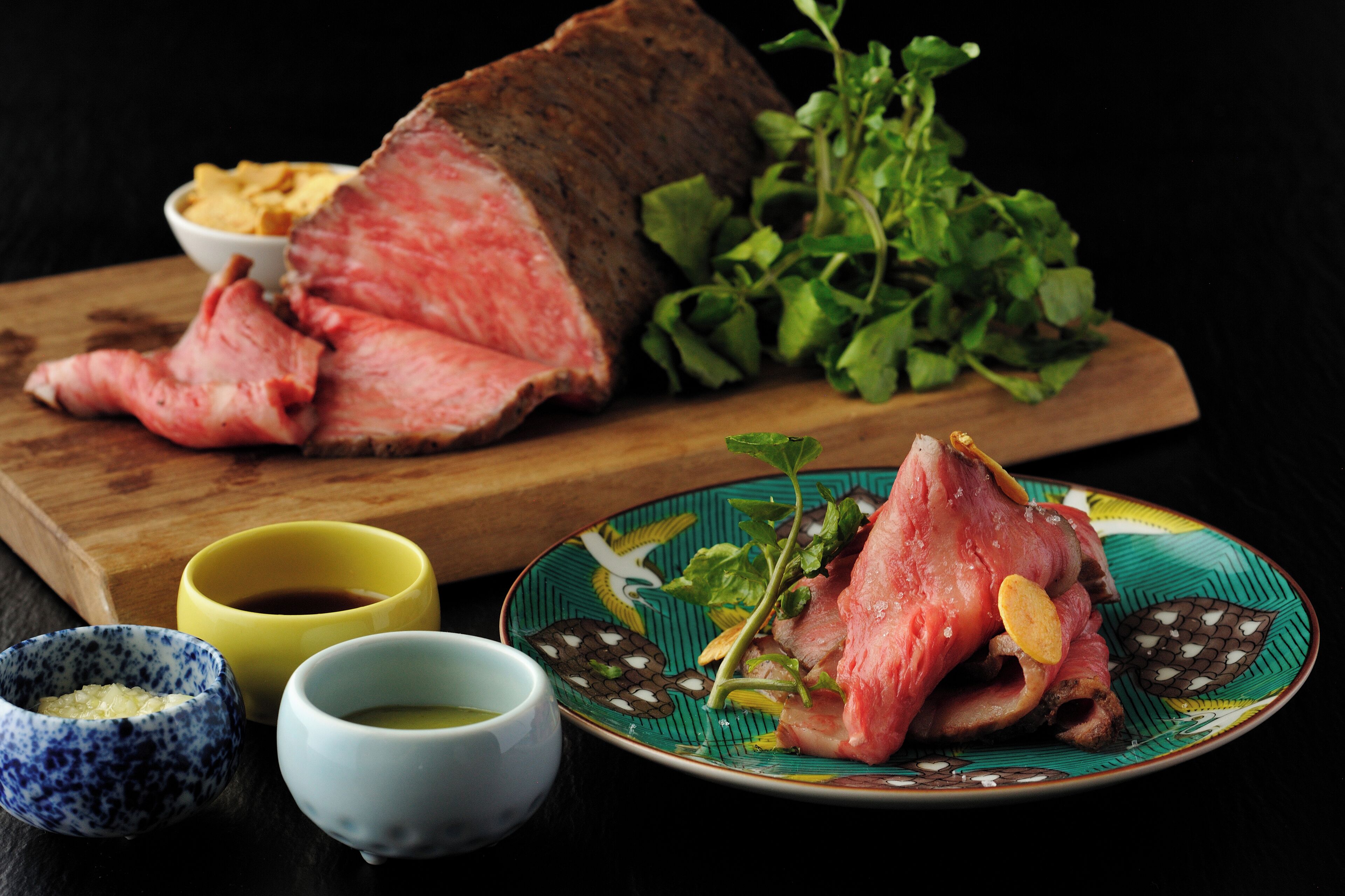 Un gros morceau de viande servi sur une planche en bois, accompagné d’un bol de frites, d’une assiette de légumes verts et de deux petits ramequins de sauces.
