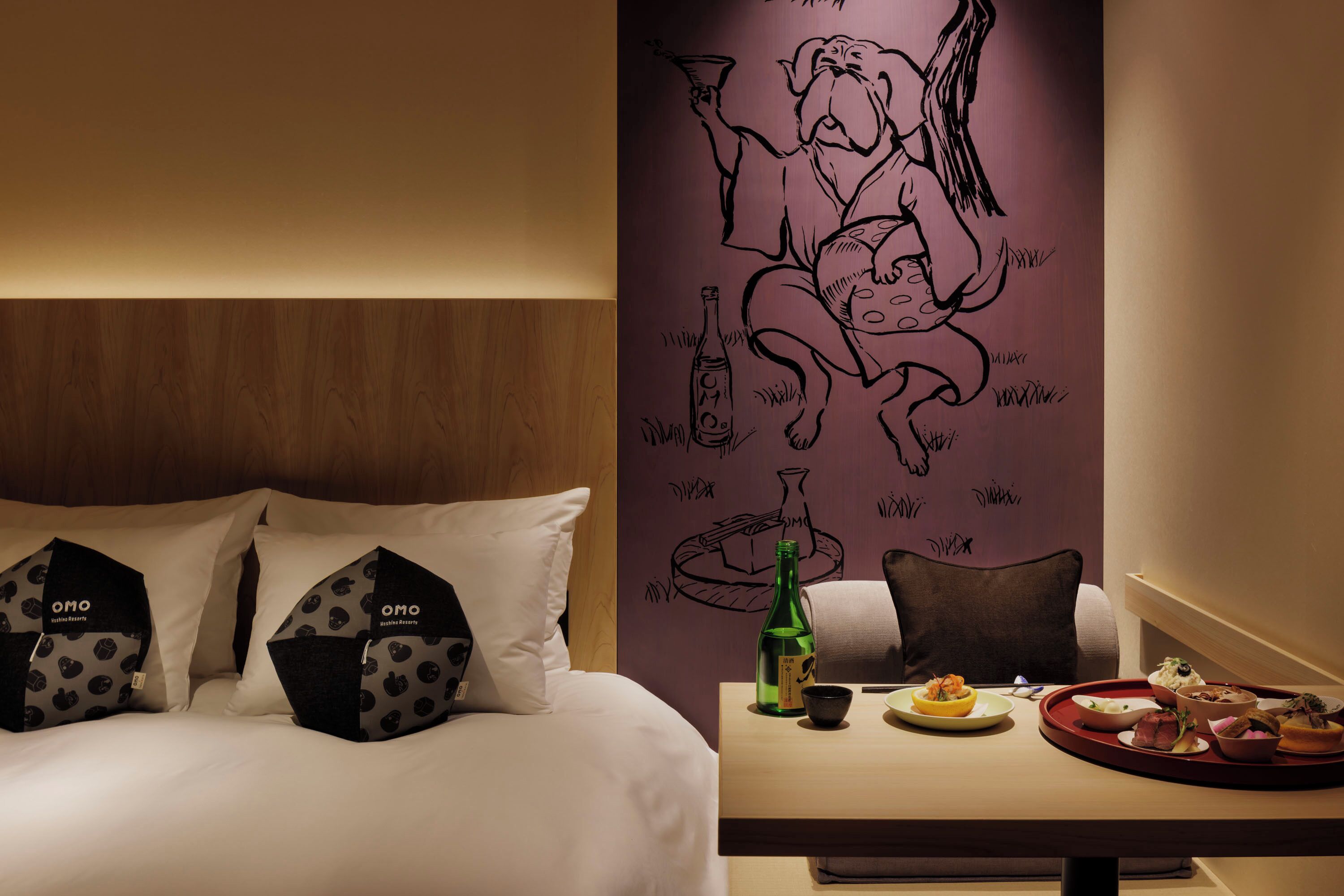 A hotel room with a bed, pillows, a table with food and drinks, and a wall with a mural.