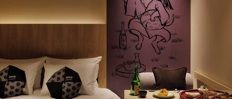 A hotel room with a bed, pillows, a table with food and drinks, and a wall with a mural.