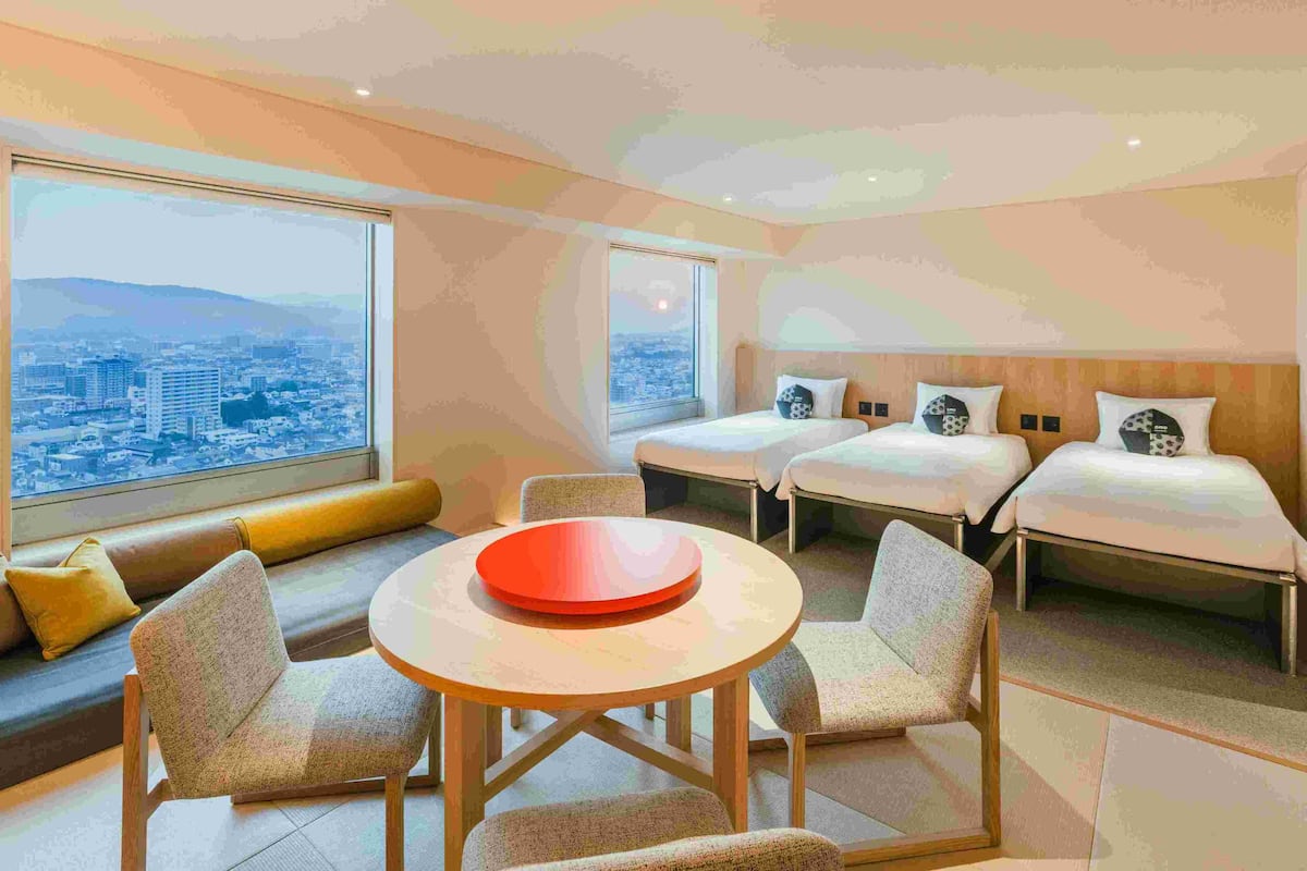 Una habitación de hotel moderna con vista a la ciudad, dos camas individuales, un sofá, una mesa de comedor y un cuenco rojo.