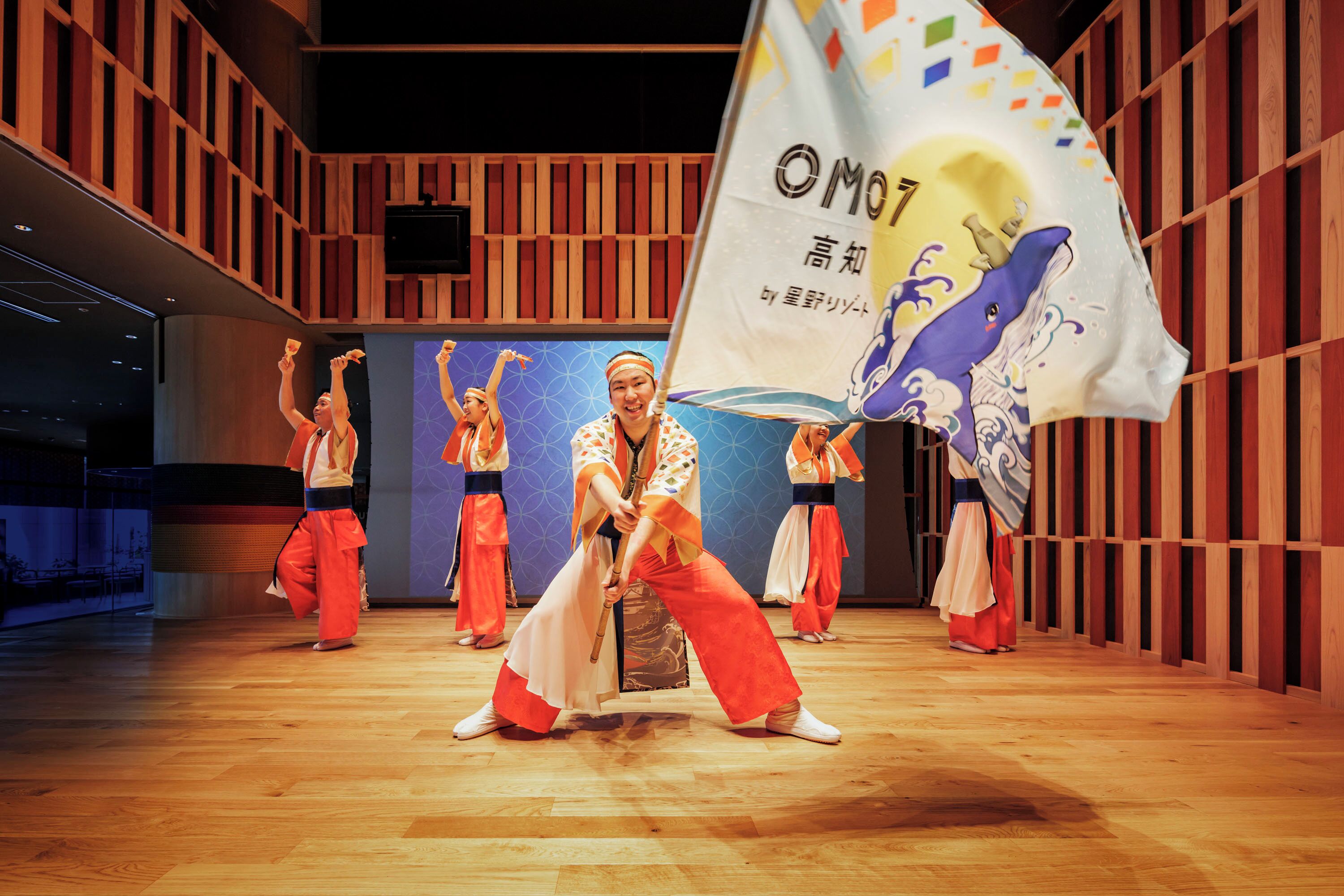 A dance performance in a wooden-paneled room with a stage and a large flag.