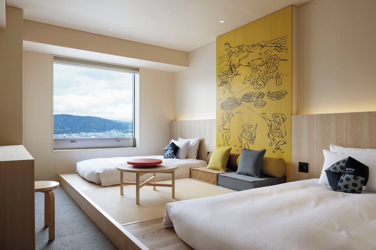 Una habitación de hotel moderna con un ventanal grande, una cama, un sofá, una mesita pequeña y una pared amarilla con dibujos negros.