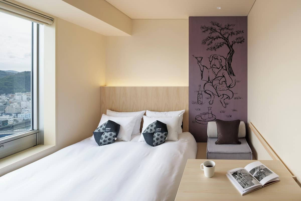 Habitación de hotel con una cama grande, vistas de la ciudad a través de una ventana, una pared con un mural de un árbol y una mesita con un libro y una taza.