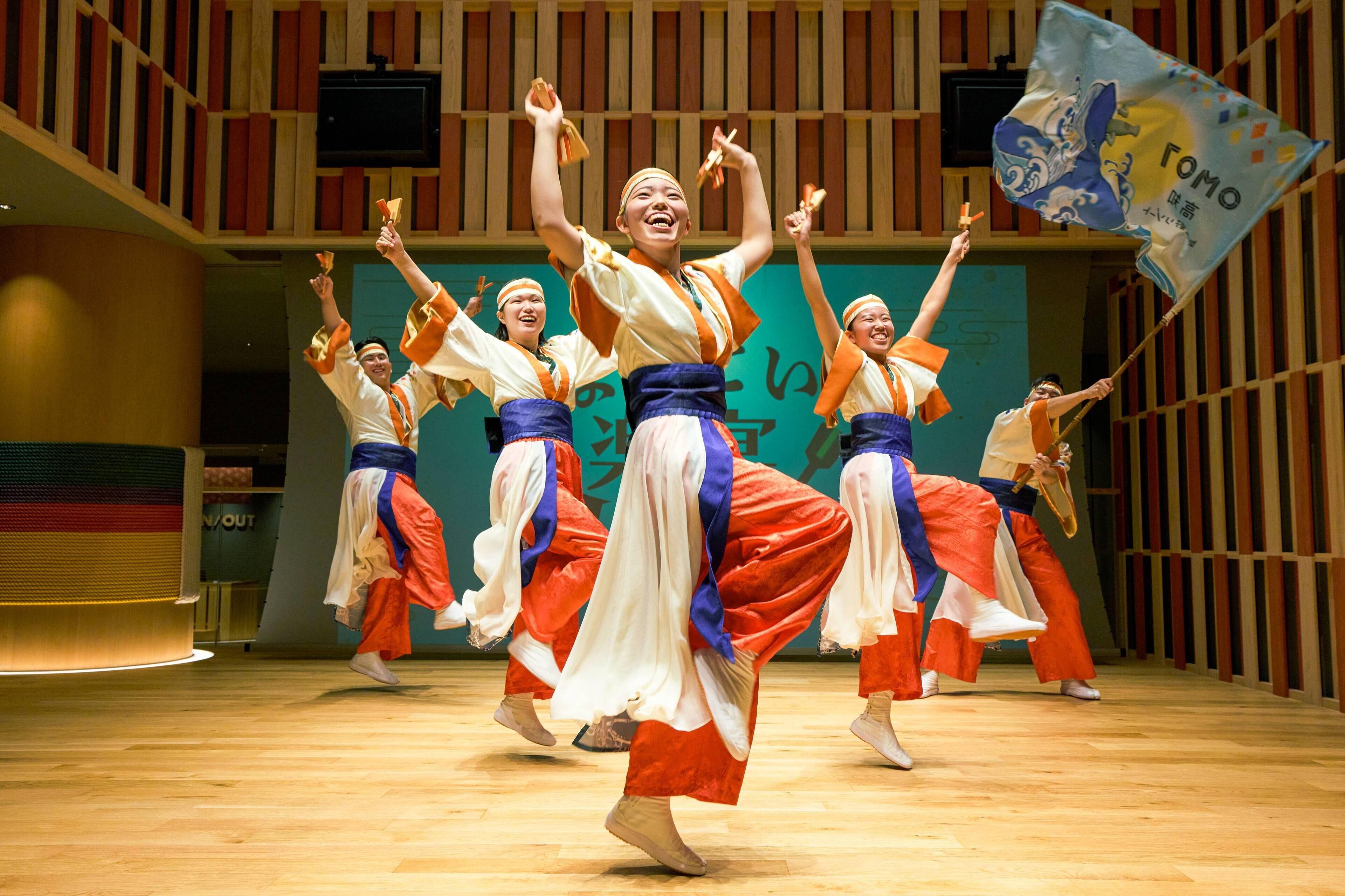 A group of dancers performing in a spacious indoor venue with a wooden floor and a large screen in the background.