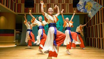 A group of dancers performing in a spacious indoor venue with a wooden floor and a large screen in the background.