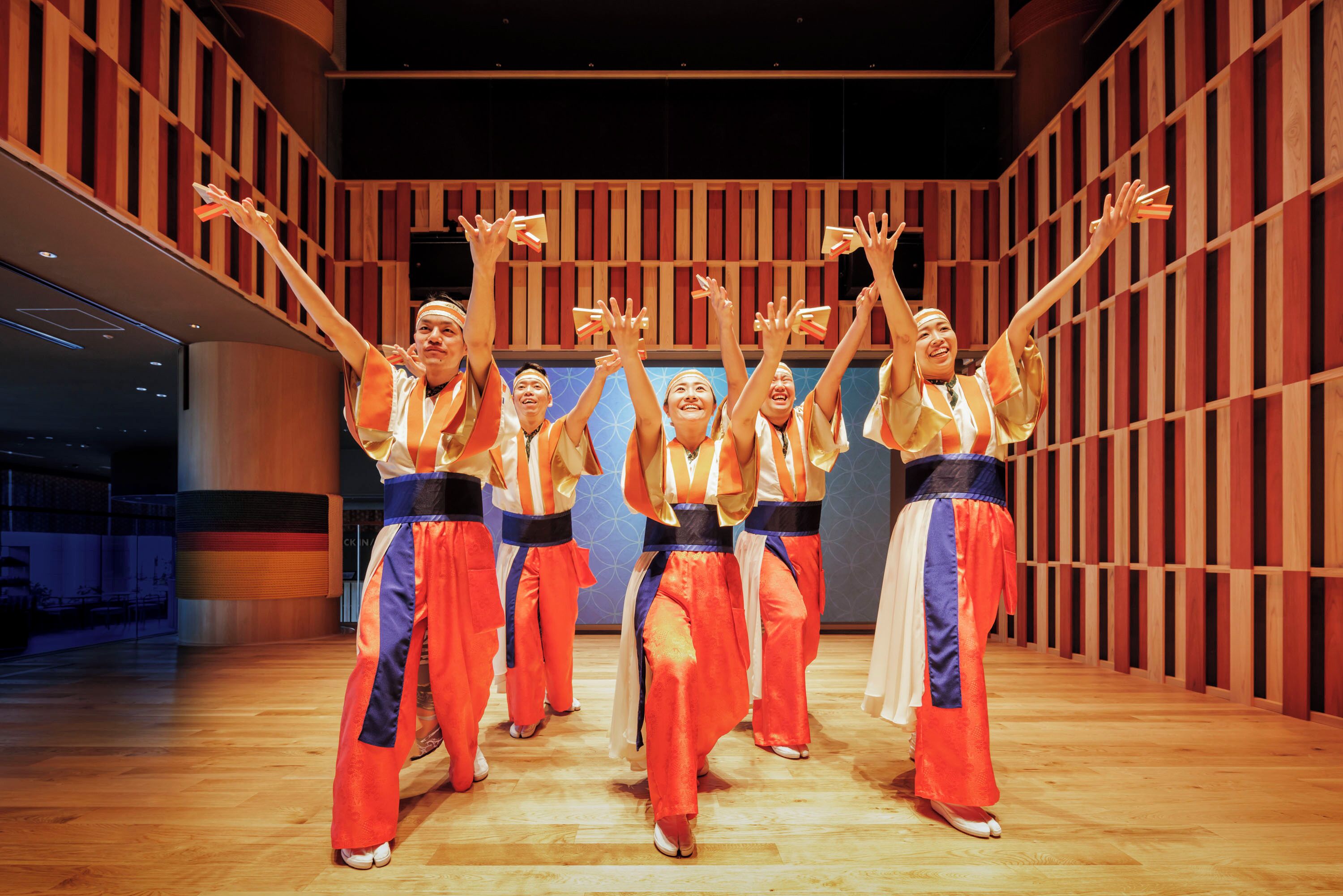 A group of people in traditional attire performing in a spacious hall with wooden paneling and a polished floor.