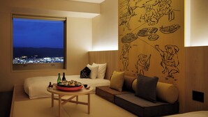A modern hotel room with a large window overlooking a cityscape at night, a cozy seating area with a sofa and a small table with a tray of food, and a wall with cultural artwork. - OMO7 Kochi by Hoshino Resorts (Kochi)