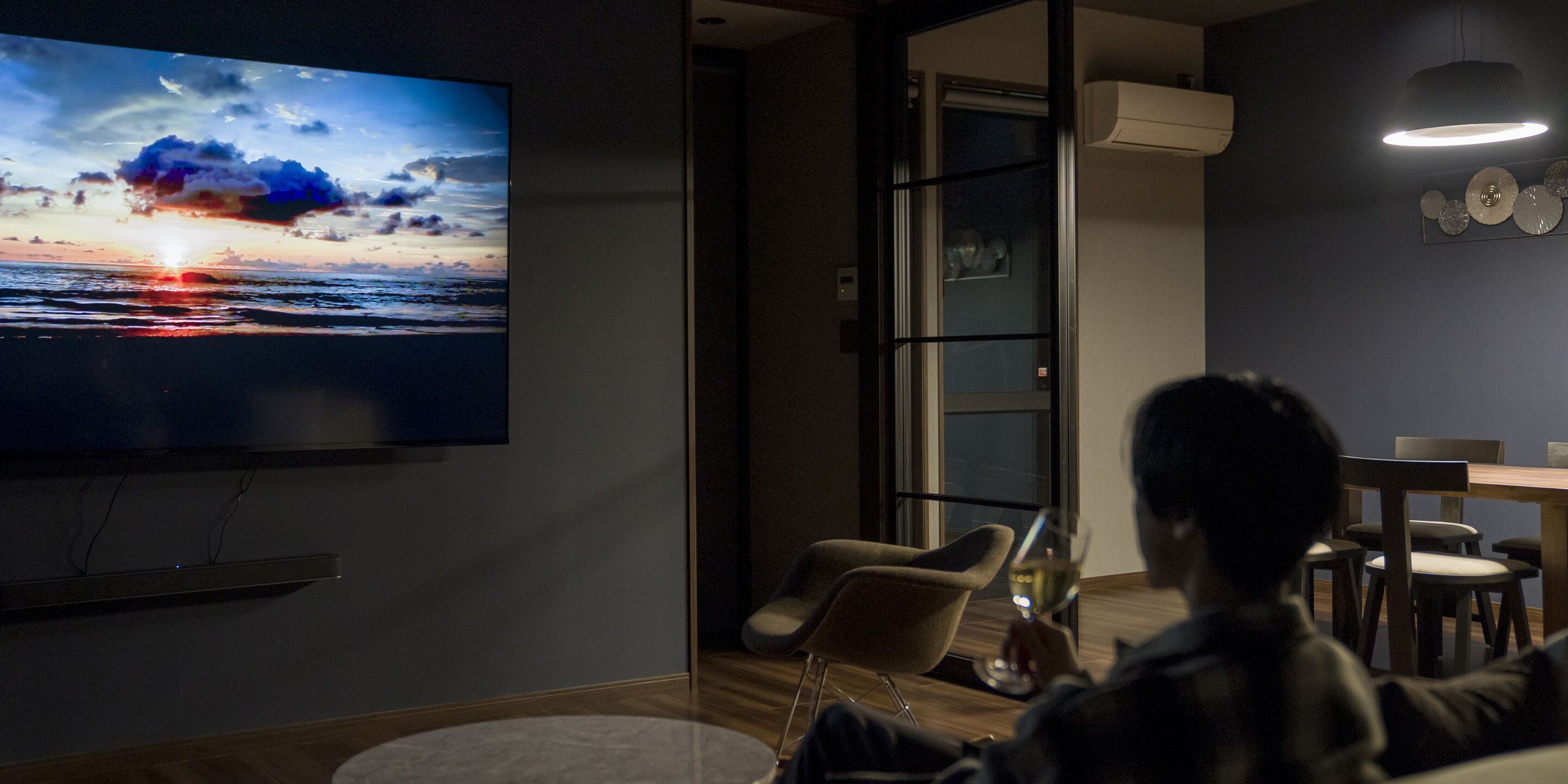Una sala moderna con un televisor de pantalla plana grande mostrando un atardecer, un comedor con mesa y sillas, y un ventanal de vidrio que da a un balcón.