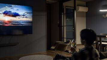 Una sala moderna con un televisor de pantalla plana grande mostrando un atardecer, un comedor con mesa y sillas, y un ventanal de vidrio que da a un balcón.