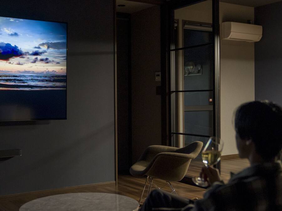 Una sala moderna con un televisor de pantalla plana grande mostrando un atardecer, un comedor con mesa y sillas, y un ventanal de vidrio que da a un balcón.