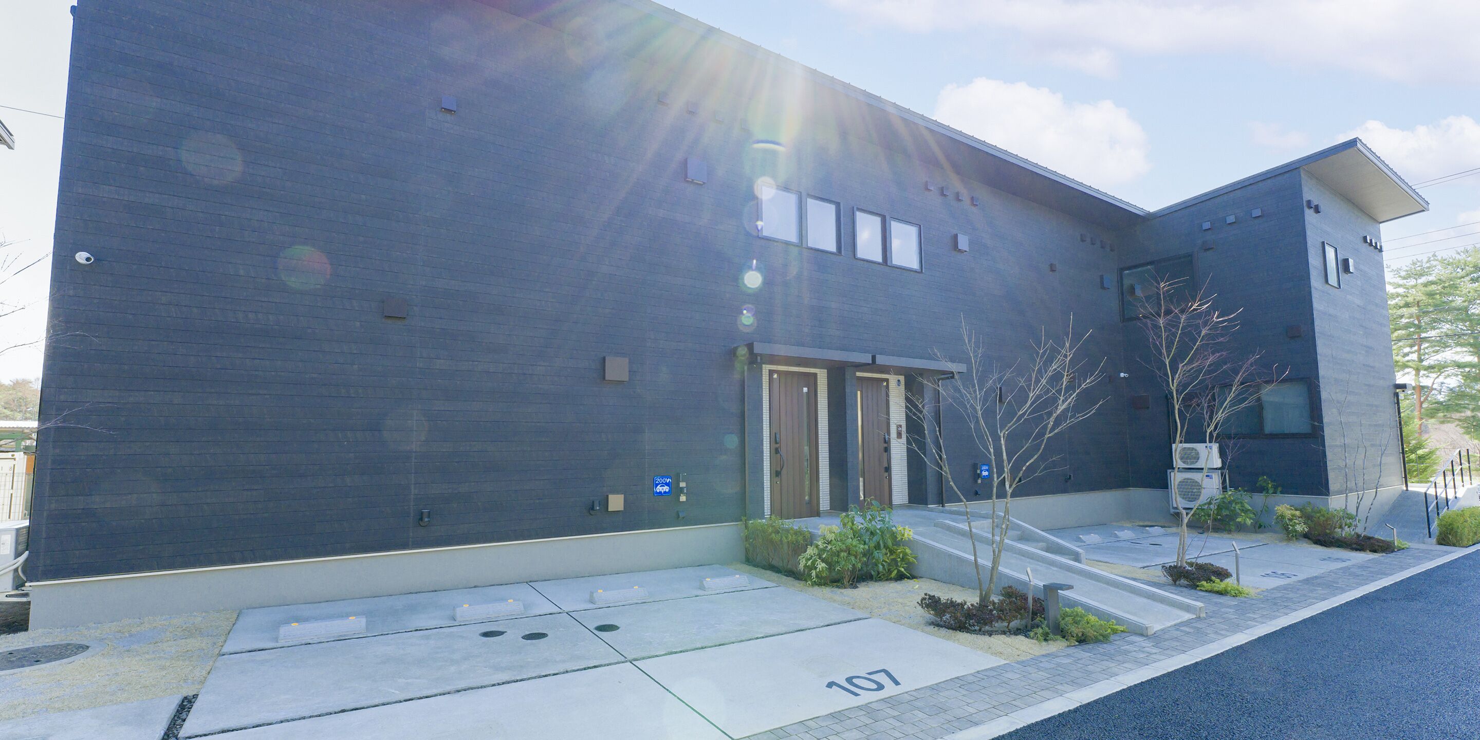 A modern building with a dark facade, a set of double doors, and a small garden area with plants and a tree.