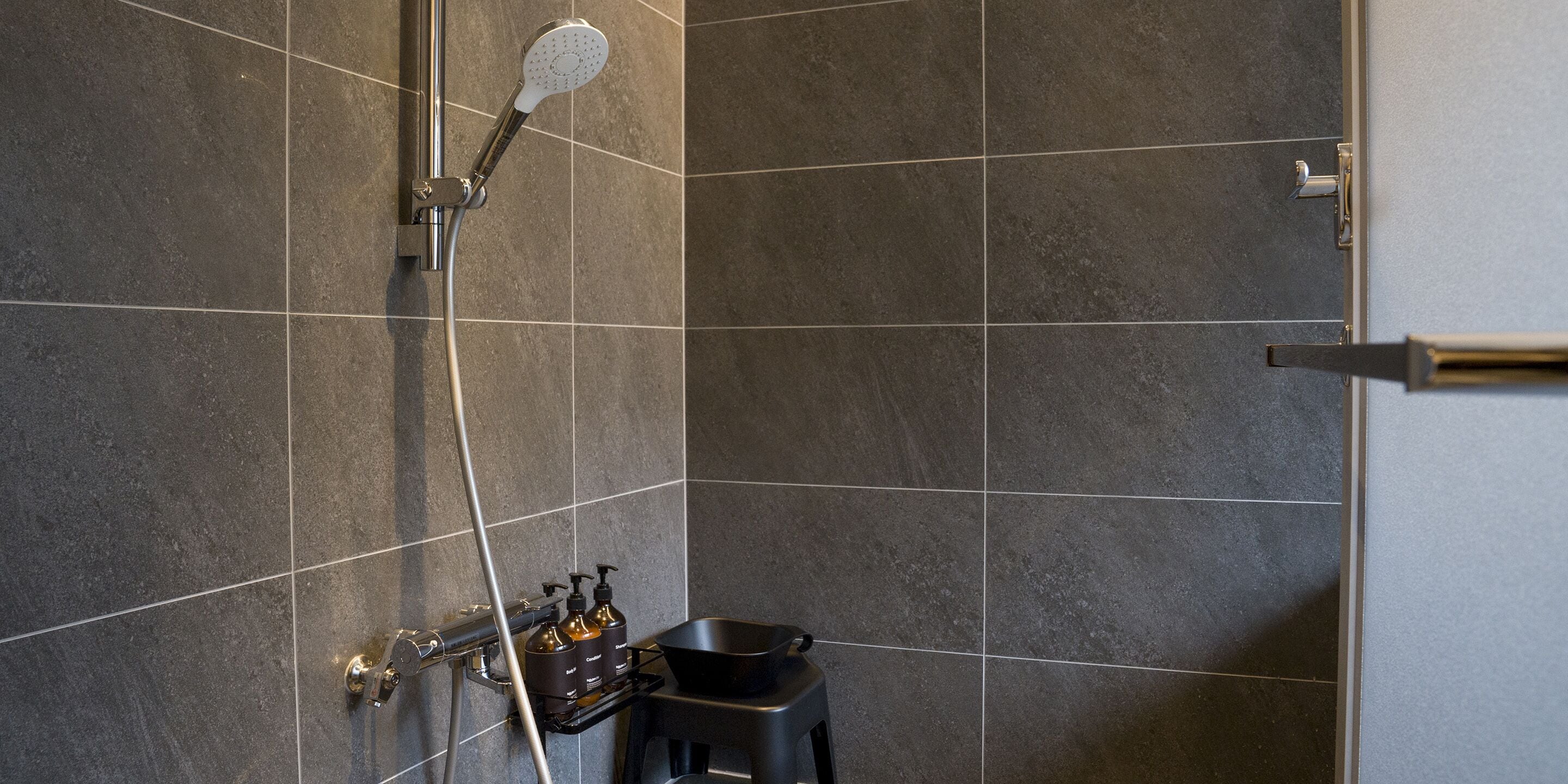 A modern bathroom with dark tiles, a showerhead, a handheld shower, and a small stool with toiletries.