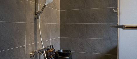 A modern bathroom with dark tiles, a showerhead, a handheld shower, and a small stool with toiletries.