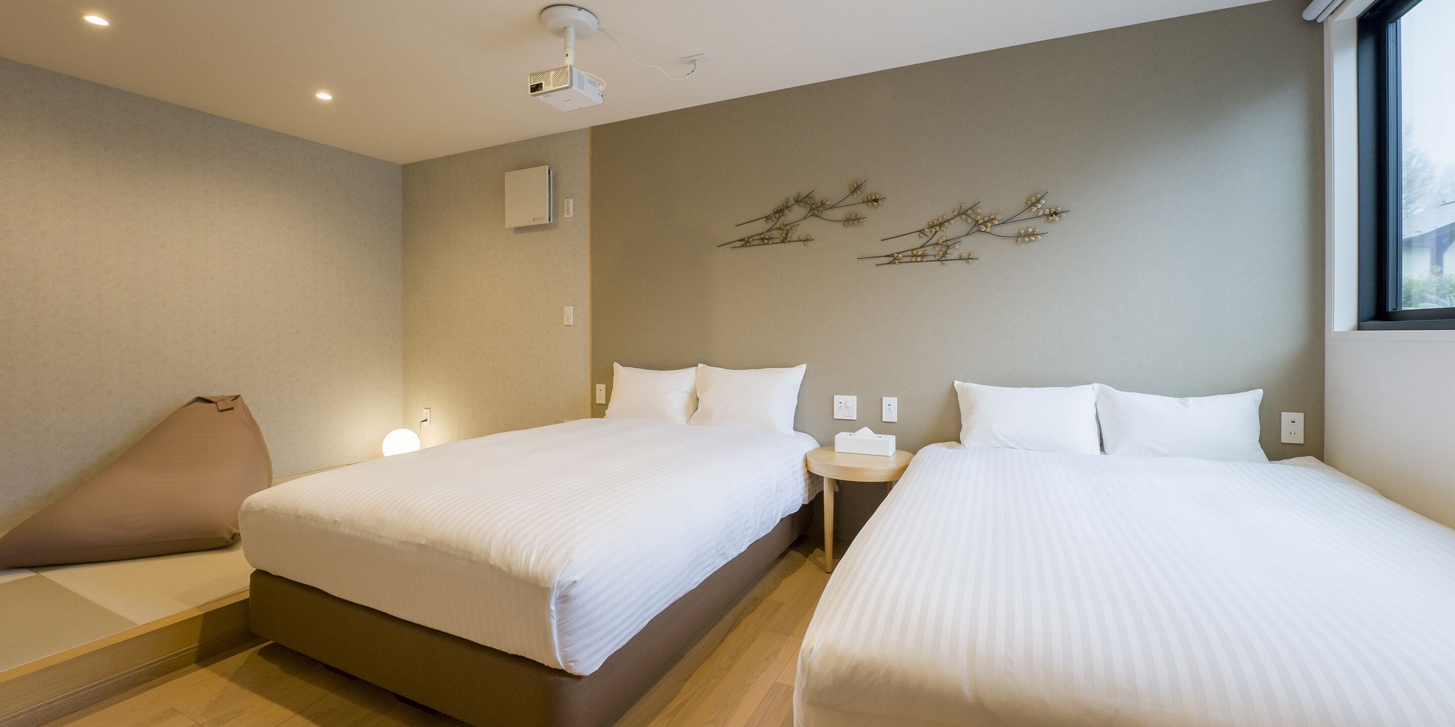 Habitación de hotel con dos camas, una mesita de noche pequeña, un proyector y aire acondicionado instalado en la pared.