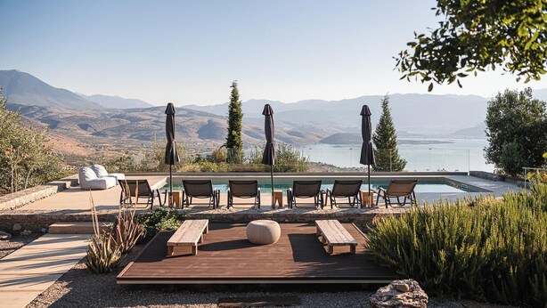 Zona junto a la piscina con sillones de descanso y sombrillas, con vistas a un lago y montañas.