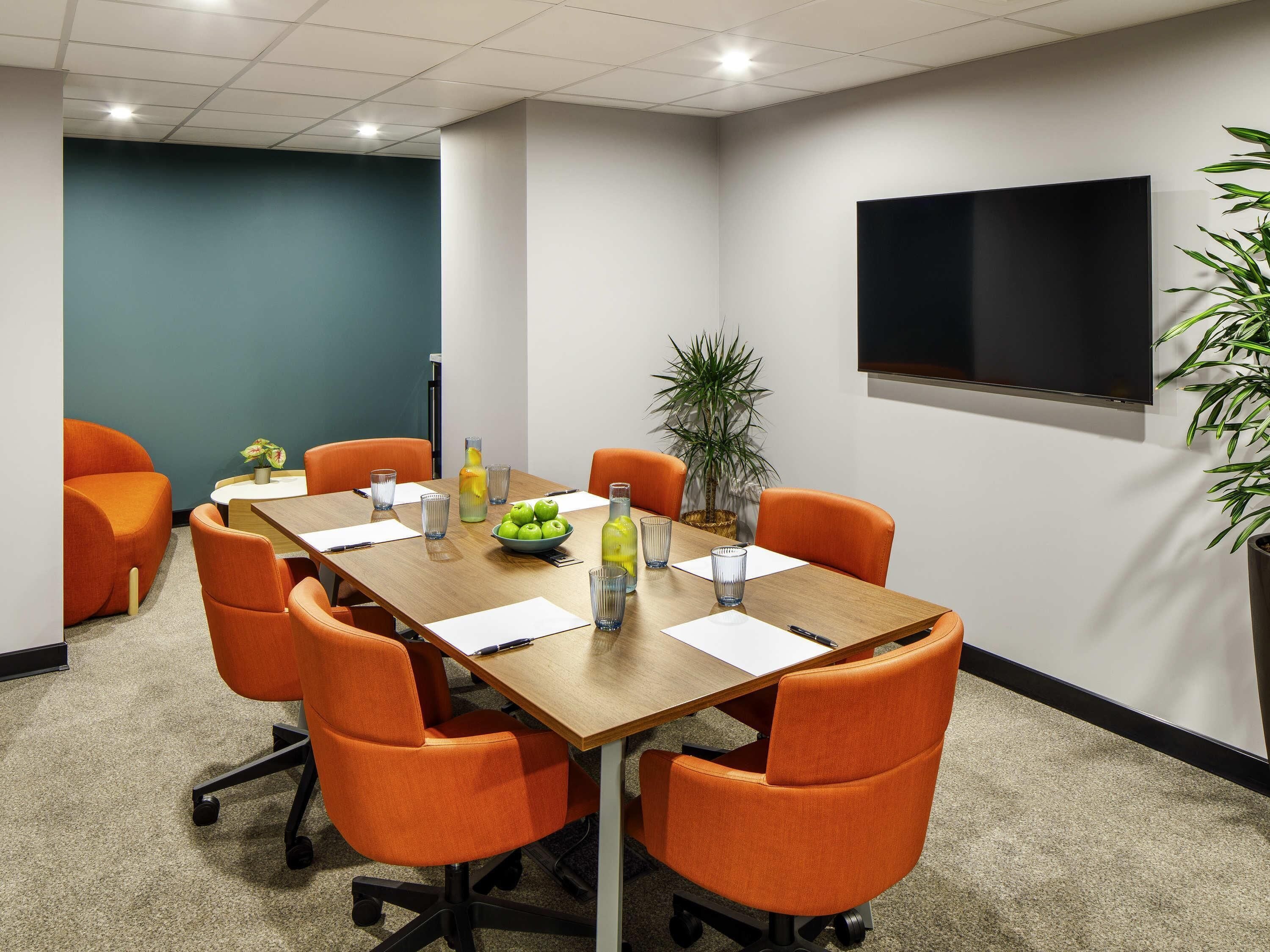A conference room with a long table, orange chairs, a TV, and a potted plant.