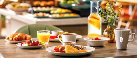 Buffet de café da manhã com diversos pratos, um jarro de suco de laranja e uma caneca sobre uma mesa de madeira.