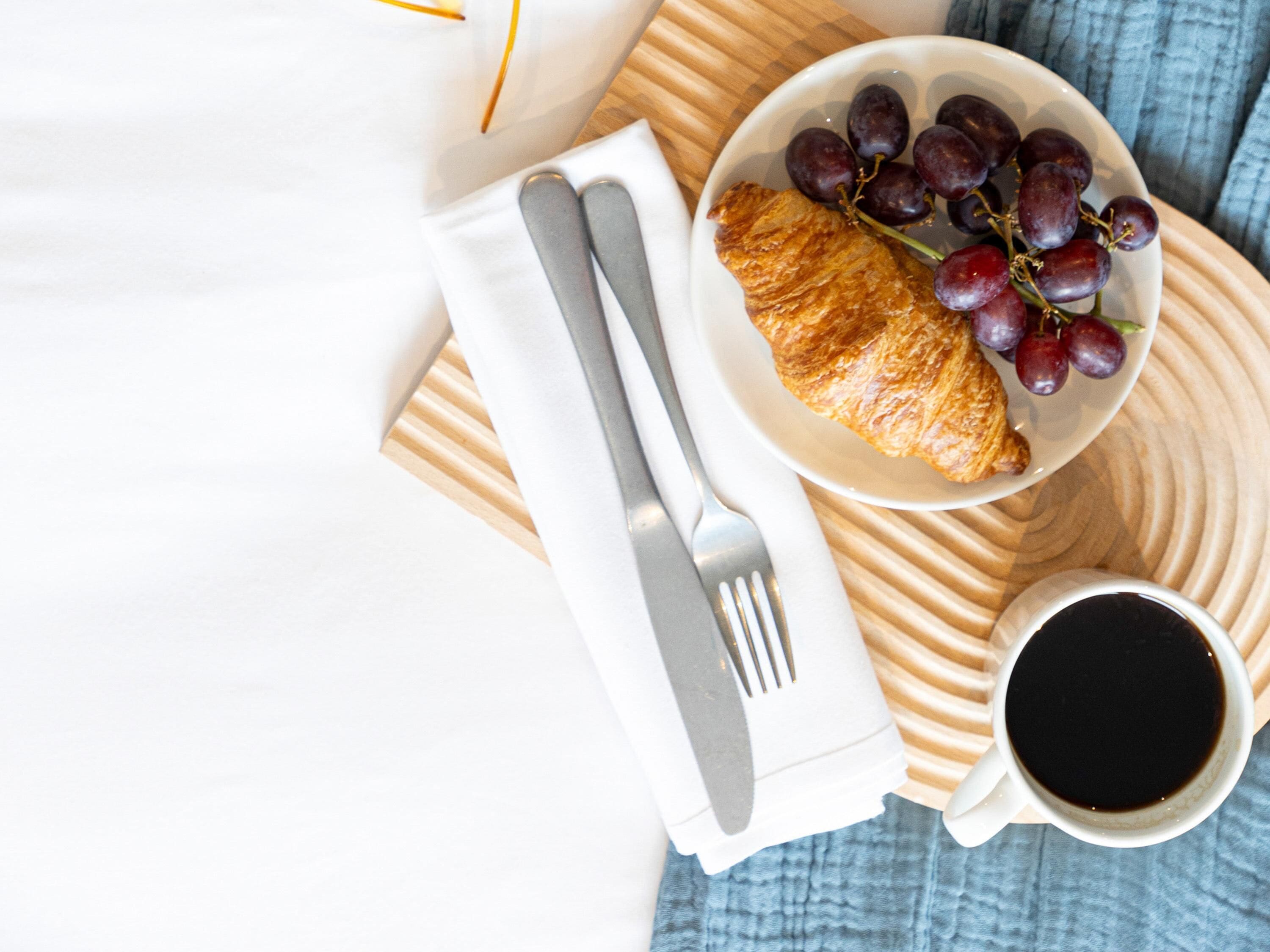 Een ontbijtje met een croissant, druiven, een kop koffie en bestek op een houten dienblad.