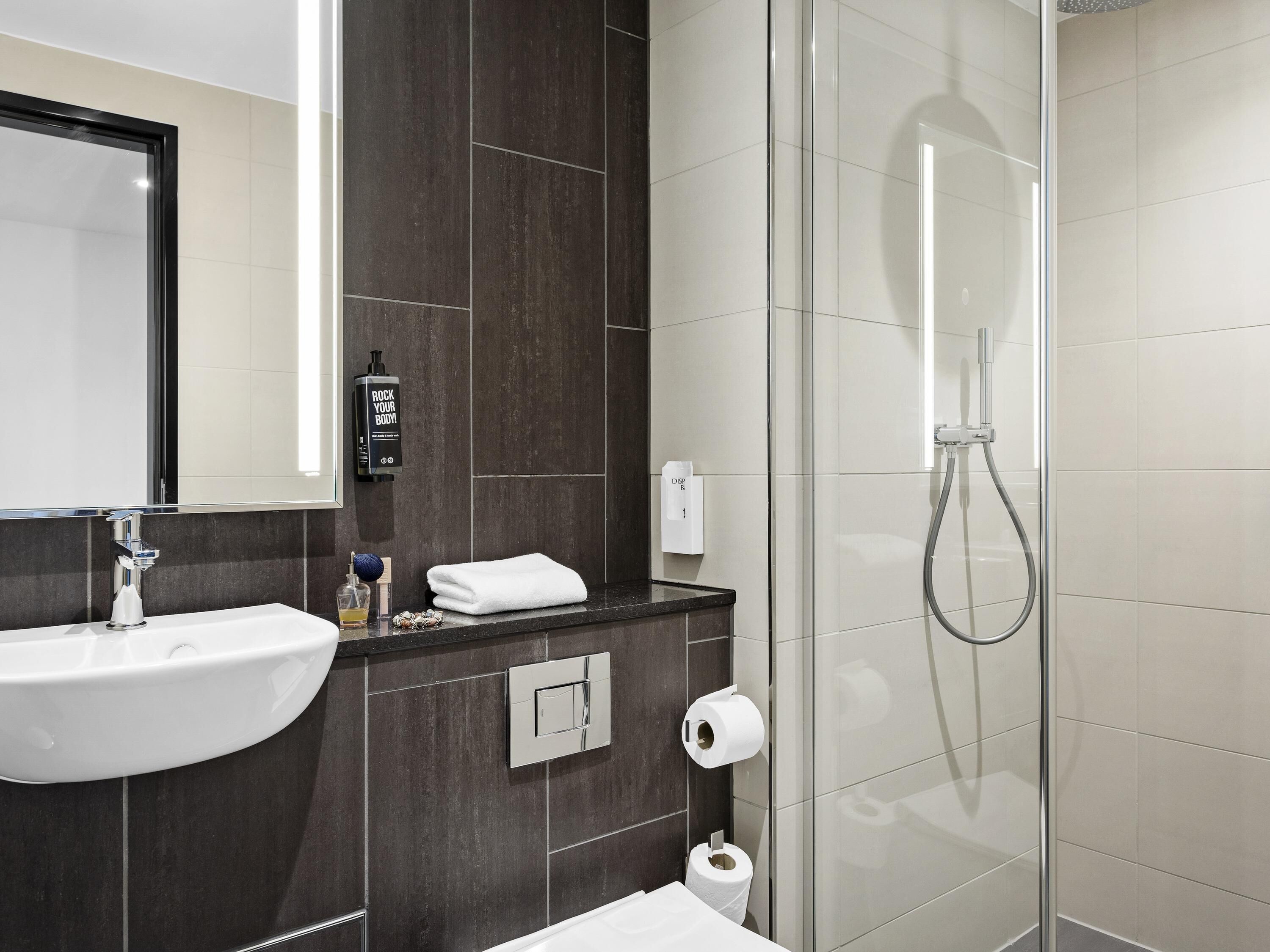 A modern bathroom with a sink, mirror, dark wall tiles, a shower enclosure, and a towel rack.