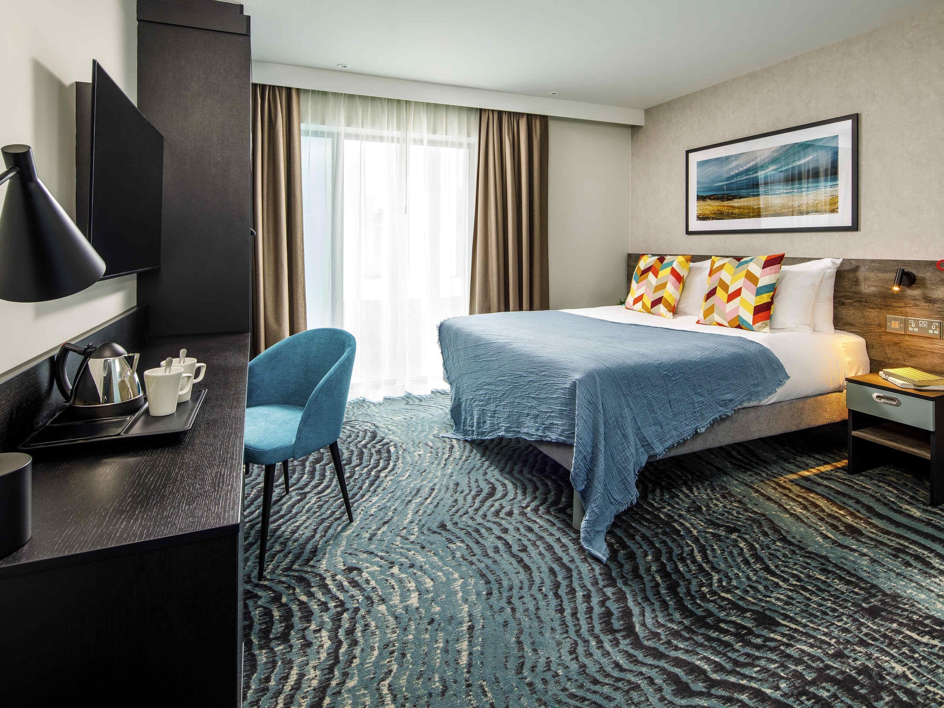 Una habitación de hotel con cama, escritorio, silla, televisión y ventana con cortinas.