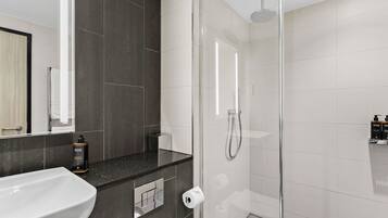 Un baño moderno con ducha envuelta en vidrio, un lavamanos blanco y paredes cubiertas de baldosas oscuras.