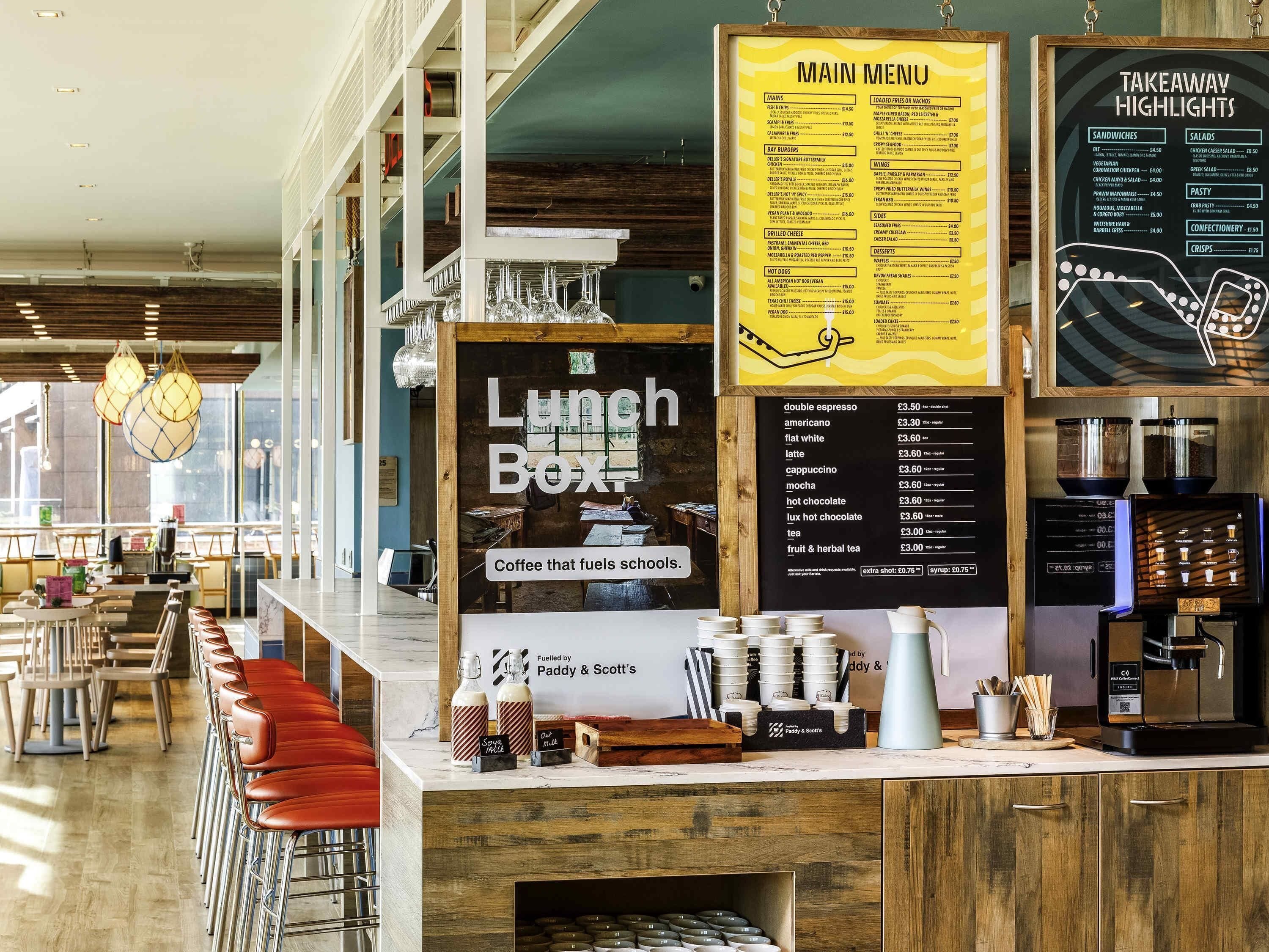 Interior de una cafetería con tablero de menú, estación de café, zona de estar y vista a la calle.