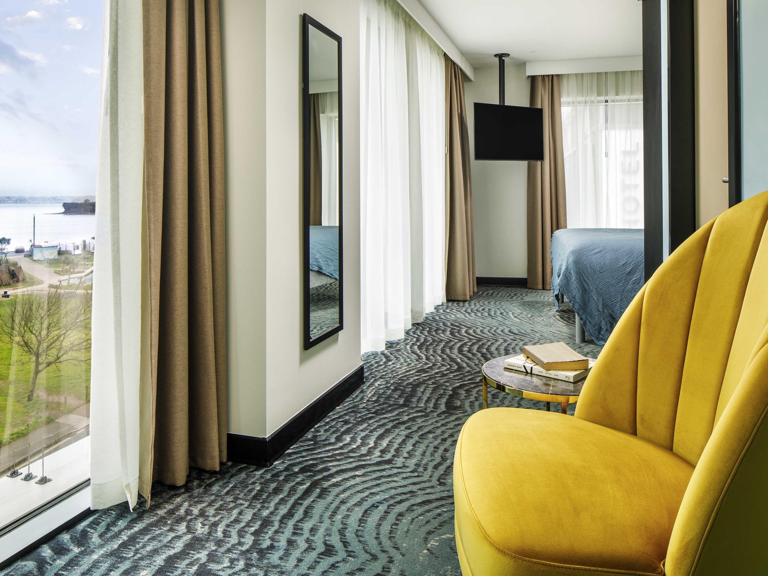 A hotel room with a bed, a yellow armchair, a flat-screen TV, and a view of a body of water.