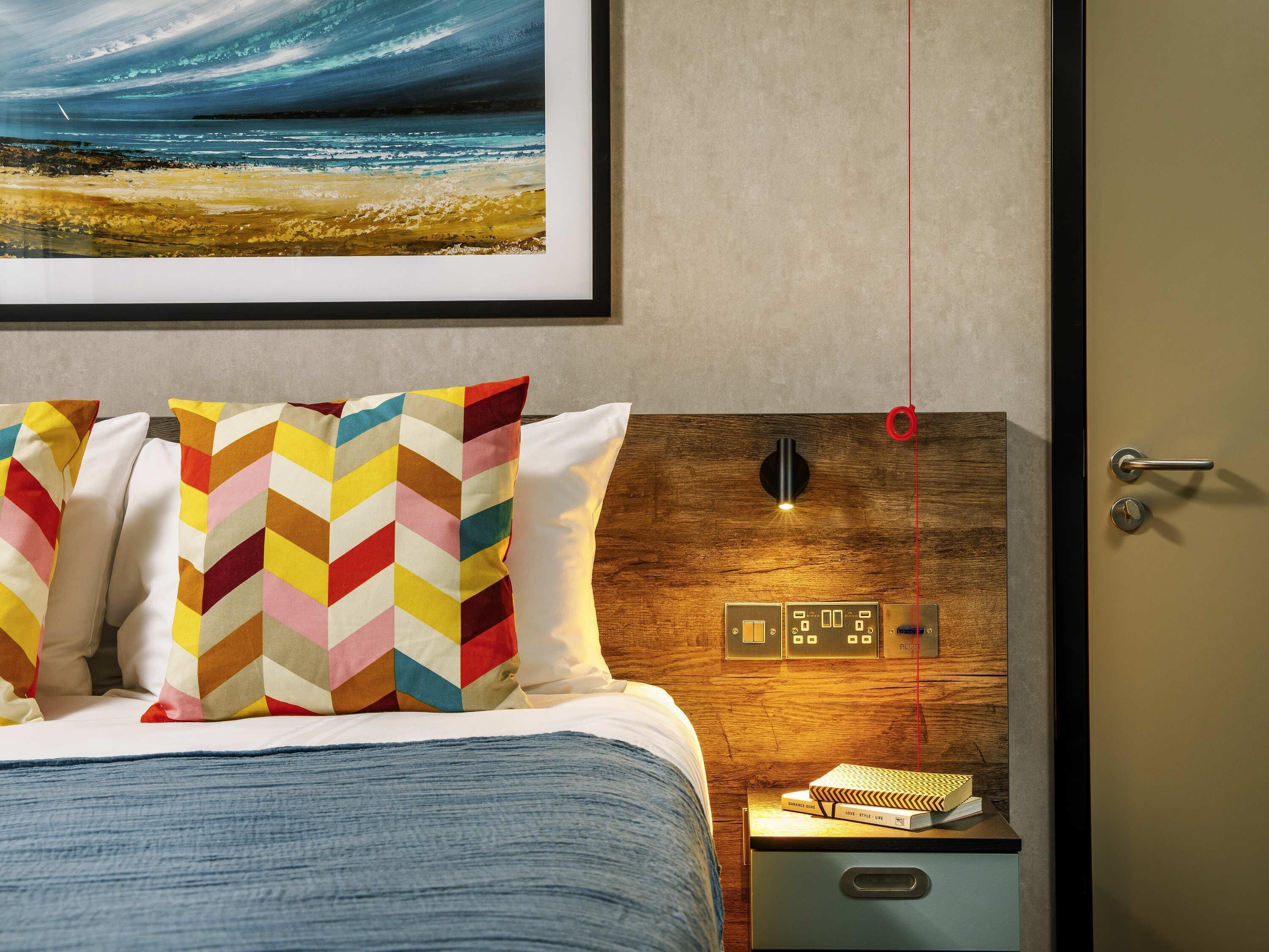 A hotel room with a bed, colorful pillows, a nightstand with books, a wall-mounted light, and a door with a lock.