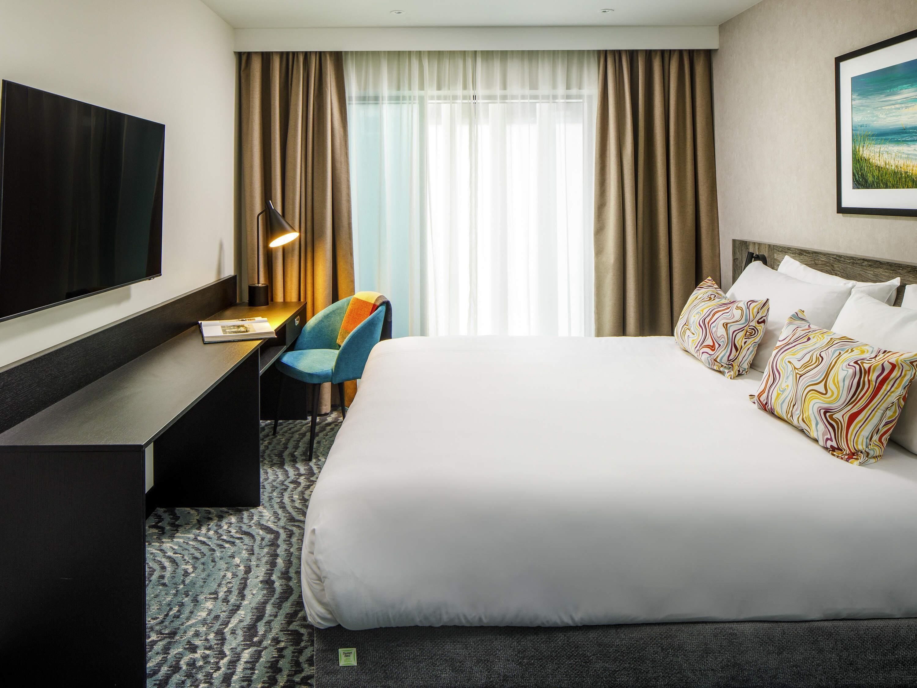 A hotel room with a large bed, a desk, a chair, a TV, and a window with curtains.