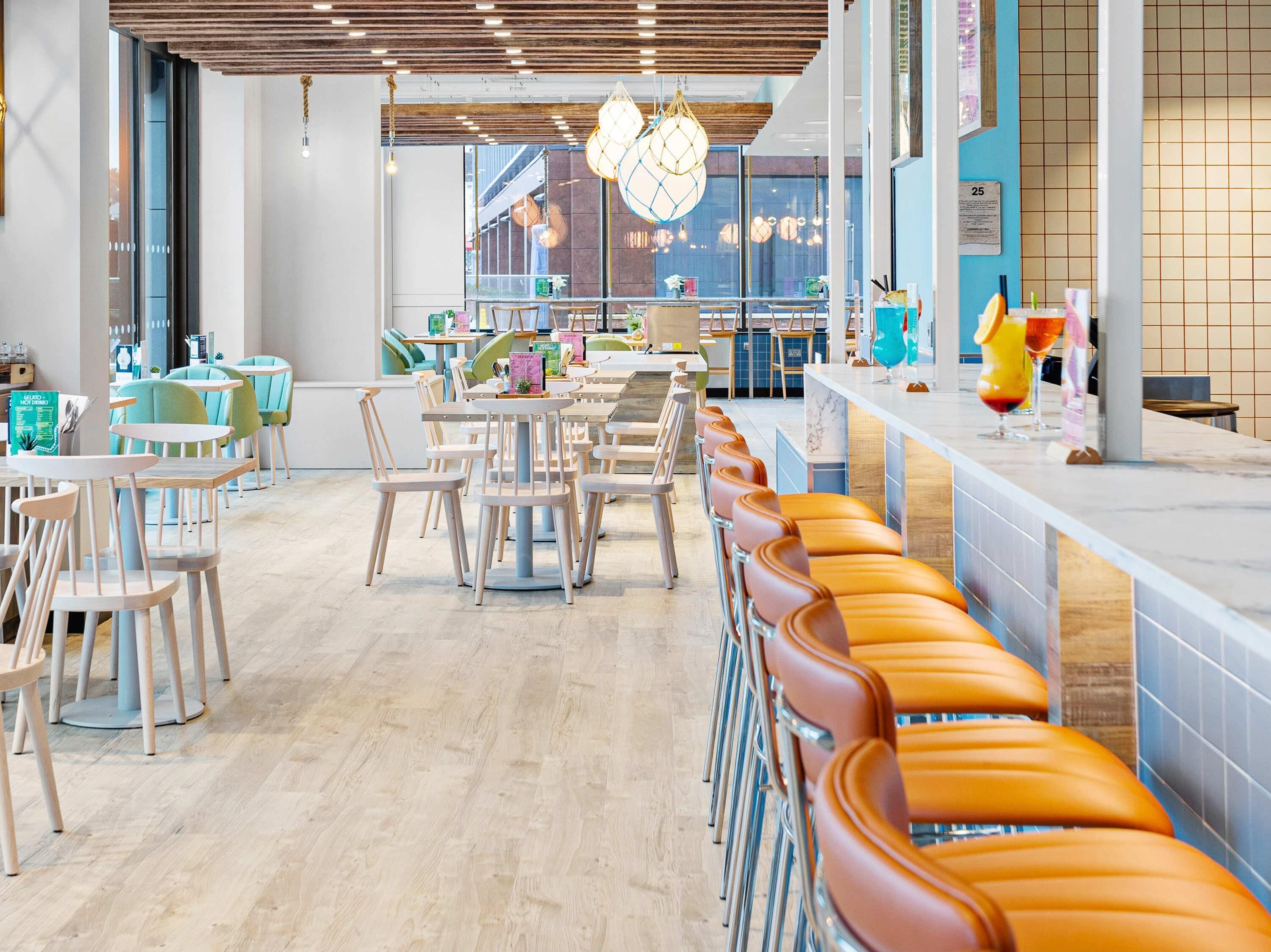 A modern cafe with wooden ceiling panels, a long counter with orange cushioned seating, and a view of the city through large windows.