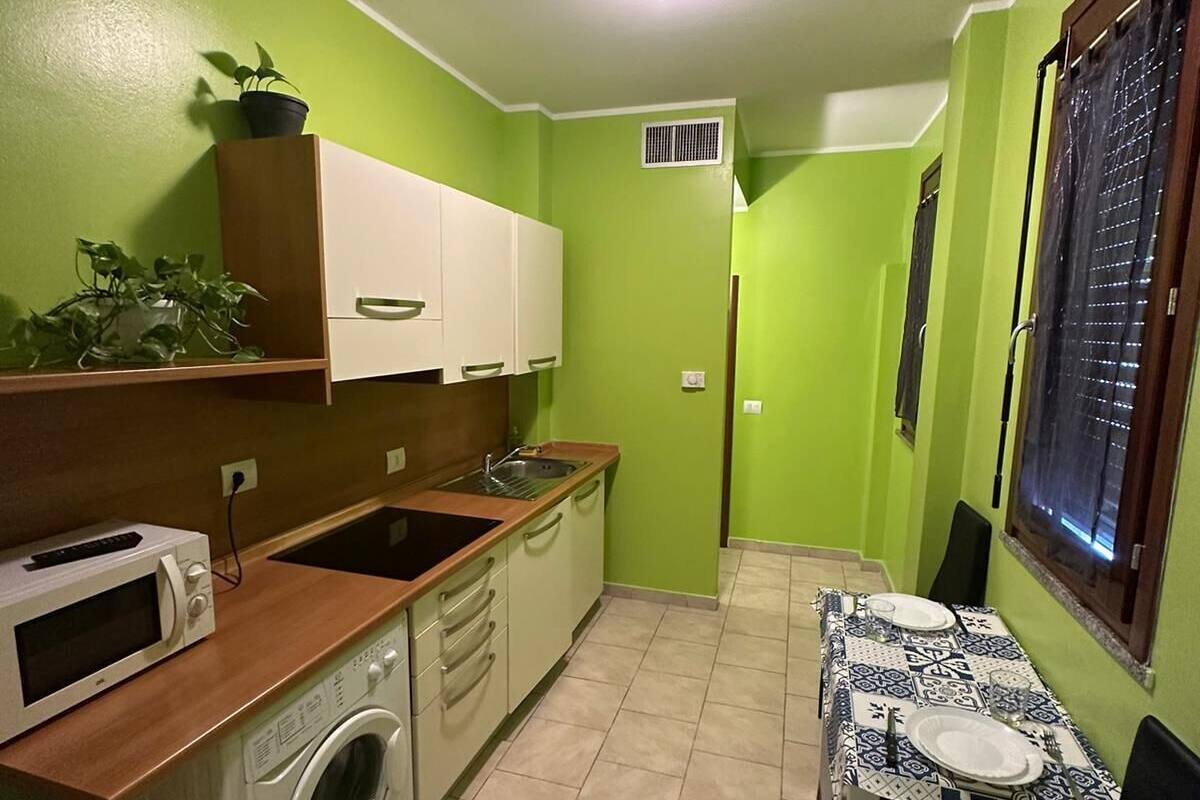 Cozinha compacta com micro-ondas, pia e máquina de lavar roupa. Há uma mesa de jantar com cadeiras e uma janela com persianas.