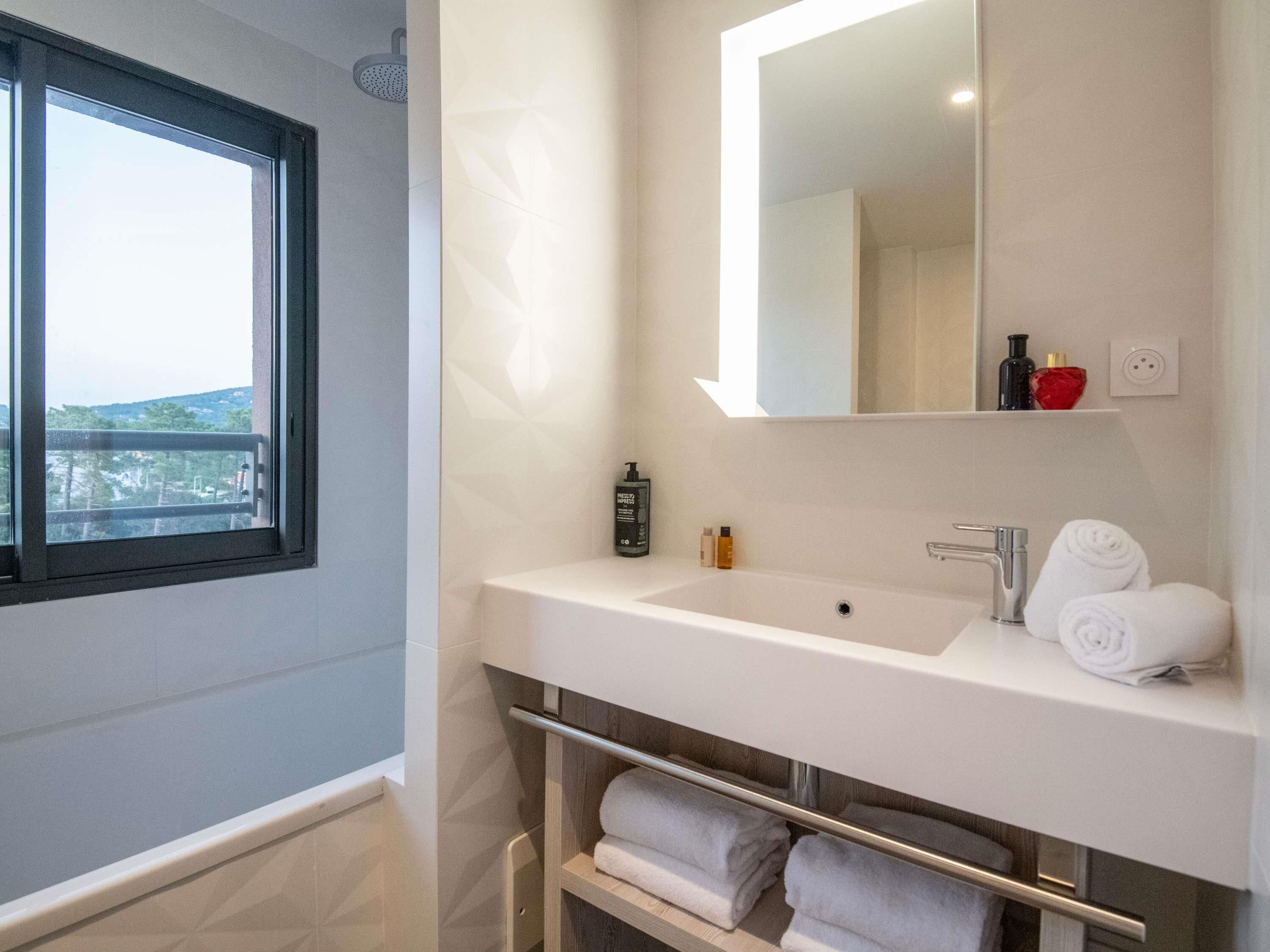A modern bathroom with a large window, a white sink, a mirror, and a towel rack.