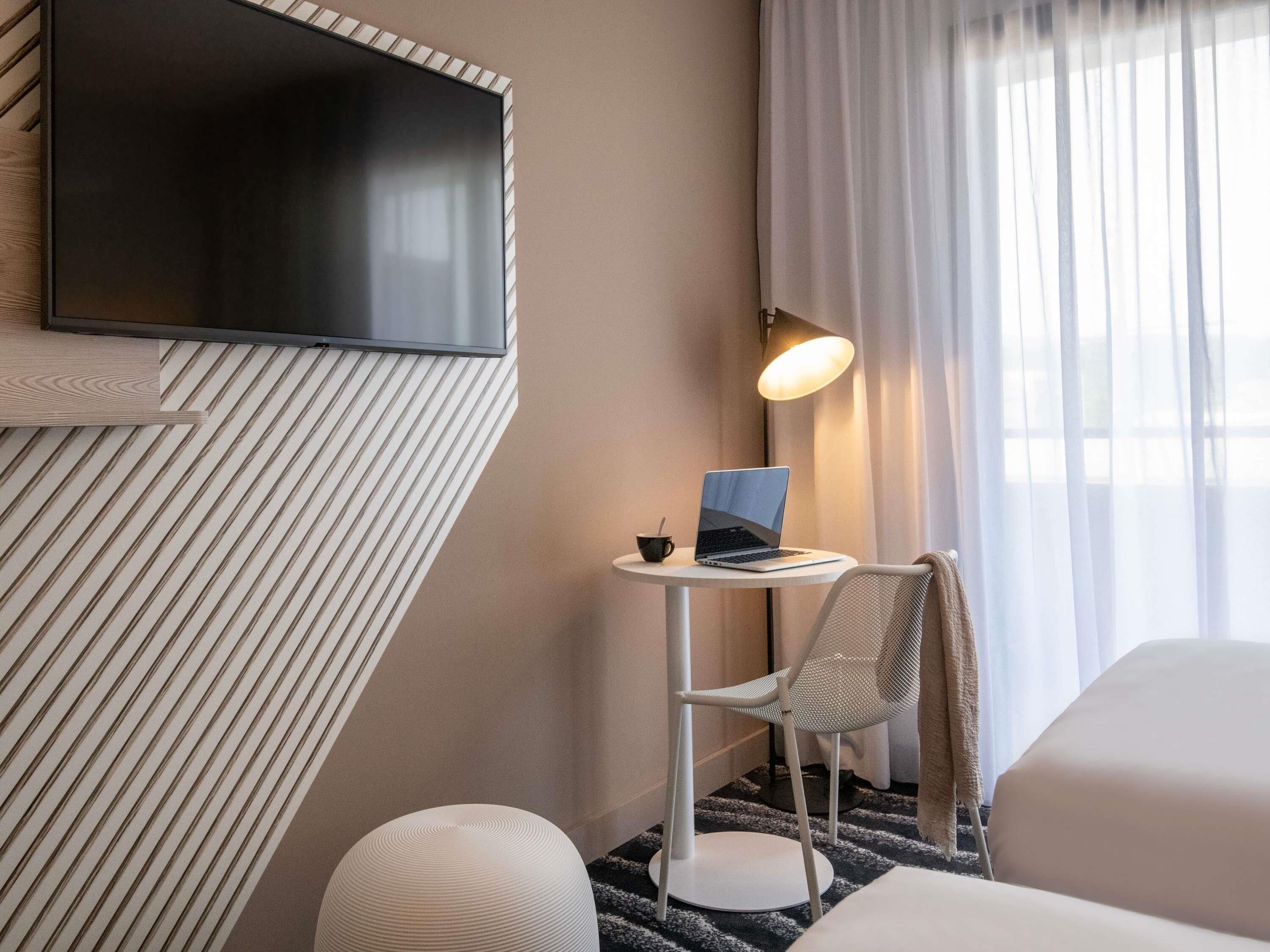 A hotel room with a flat-screen TV mounted on the wall, a small round table with a laptop, a white chair, and a bed with a beige blanket.