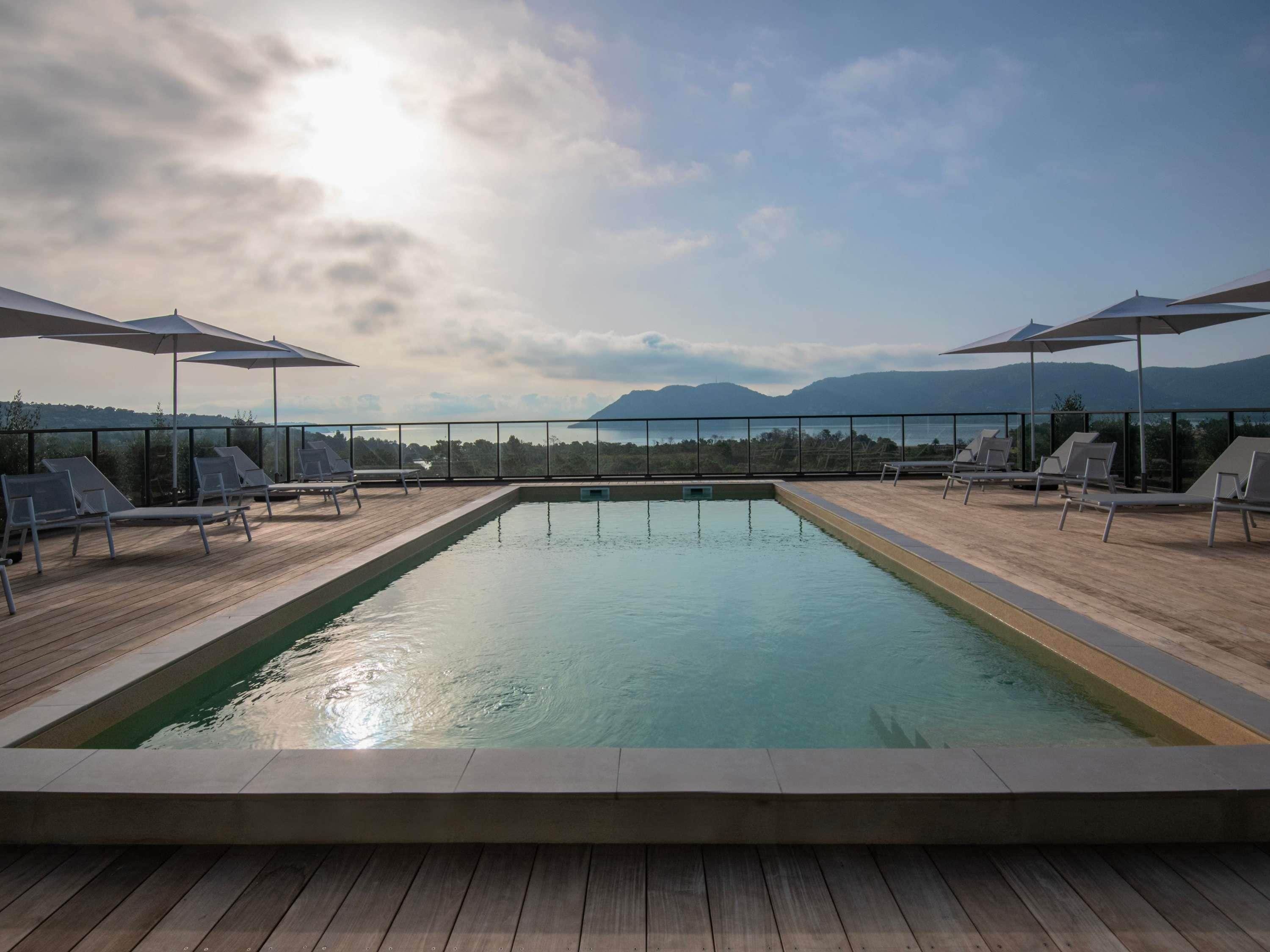 A rectangular pool with a wooden deck, surrounded by lounge chairs and umbrellas, overlooking a mountainous landscape.