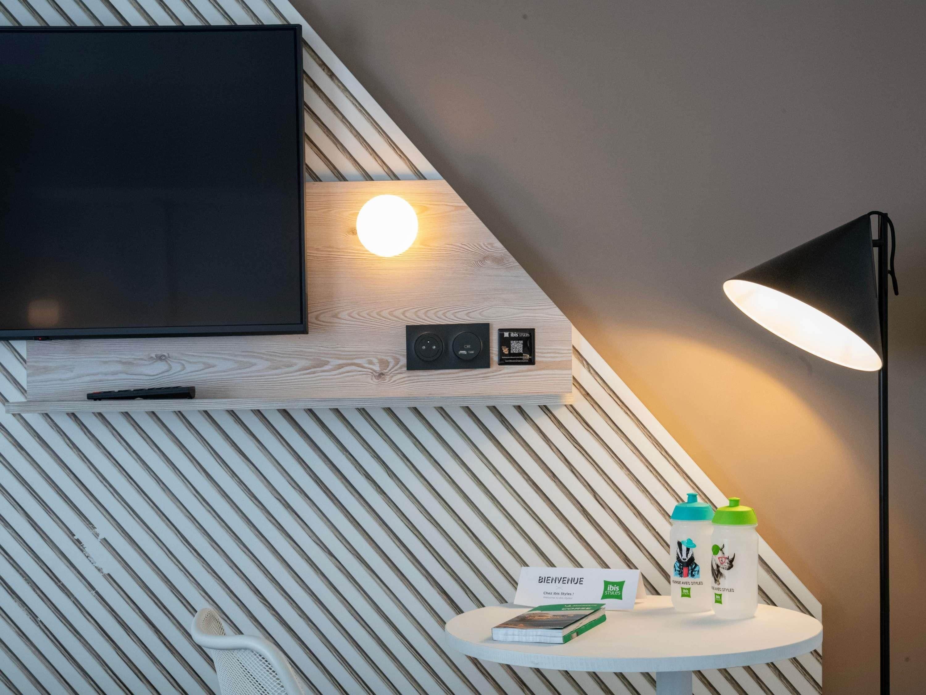 A modern room with a mounted TV, a round wall light, a table with a book and bottles, and a 'BIENVENUE' sign.