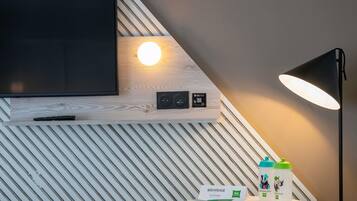 Una habitación moderna con un televisor montado, una lámpara de pared redonda, una mesa con un libro y botellas, y un cartel que dice 'BIENVENUE'.