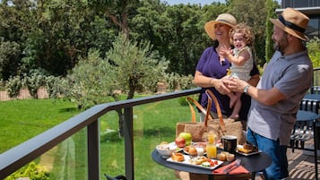 Una familia disfrutando de una comida al aire libre, con una mesa llena de comida y bebidas, rodeada de follaje y una barandilla.