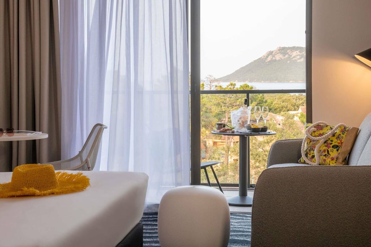 A hotel room with a bed, a chair, a small table with a bottle and glasses, and a view of a mountain and sea.