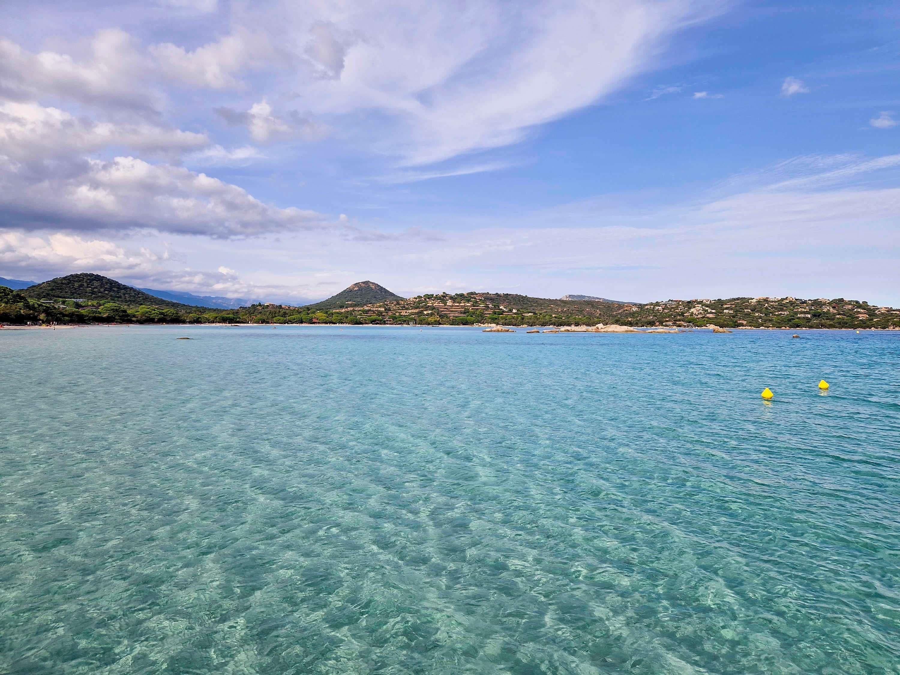 Une vue sur la côte, avec une eau limpide, une montagne lointaine et une rive bordée de bâtiments.