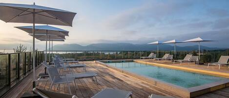 Zona de piscina con sillones de descanso y sombrillas, con vista a un cuerpo de agua y montañas lejanas.
