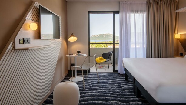 A hotel room with a bed, a small table with a chair, a round ottoman, a mirror, and a view of the sea through a balcony with curtains. - Ibis Styles Porto Vecchio (Porto-Vecchio)