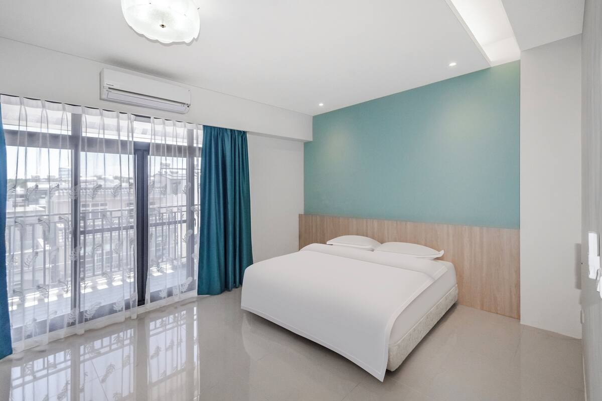 A modern hotel room with a large bed, a view of the city through floor-to-ceiling windows, and a teal accent wall.