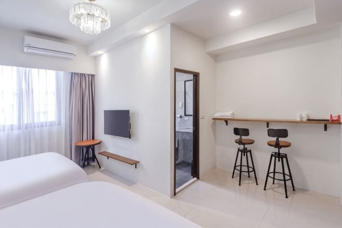 A hotel room with a bed, a TV, a desk, two stools, a shelf, and a door leading to a bathroom.