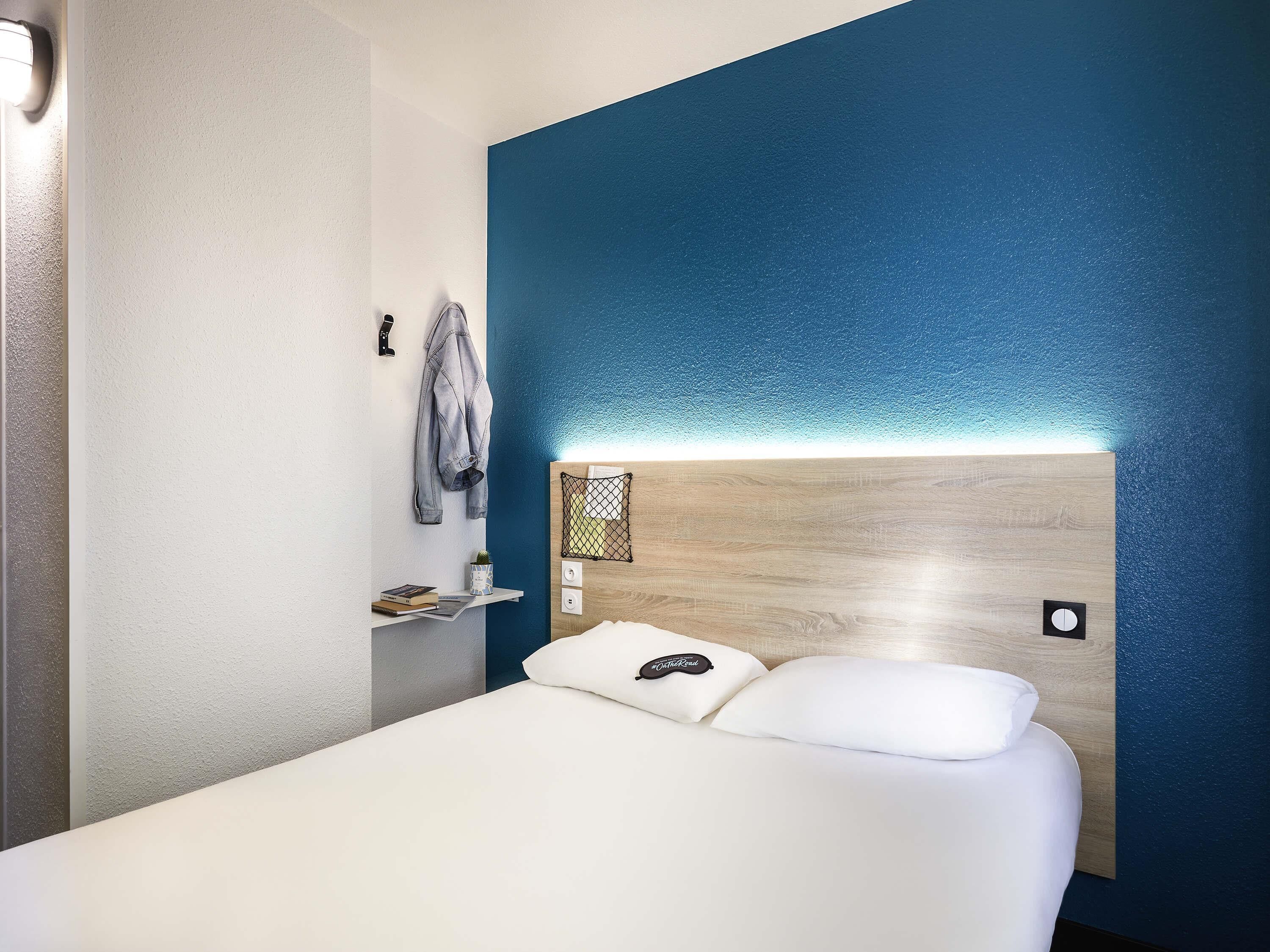 Habitación de hotel con cama, cabecera con luz, repisa con un libro y una planta, y pared con detalle azul.