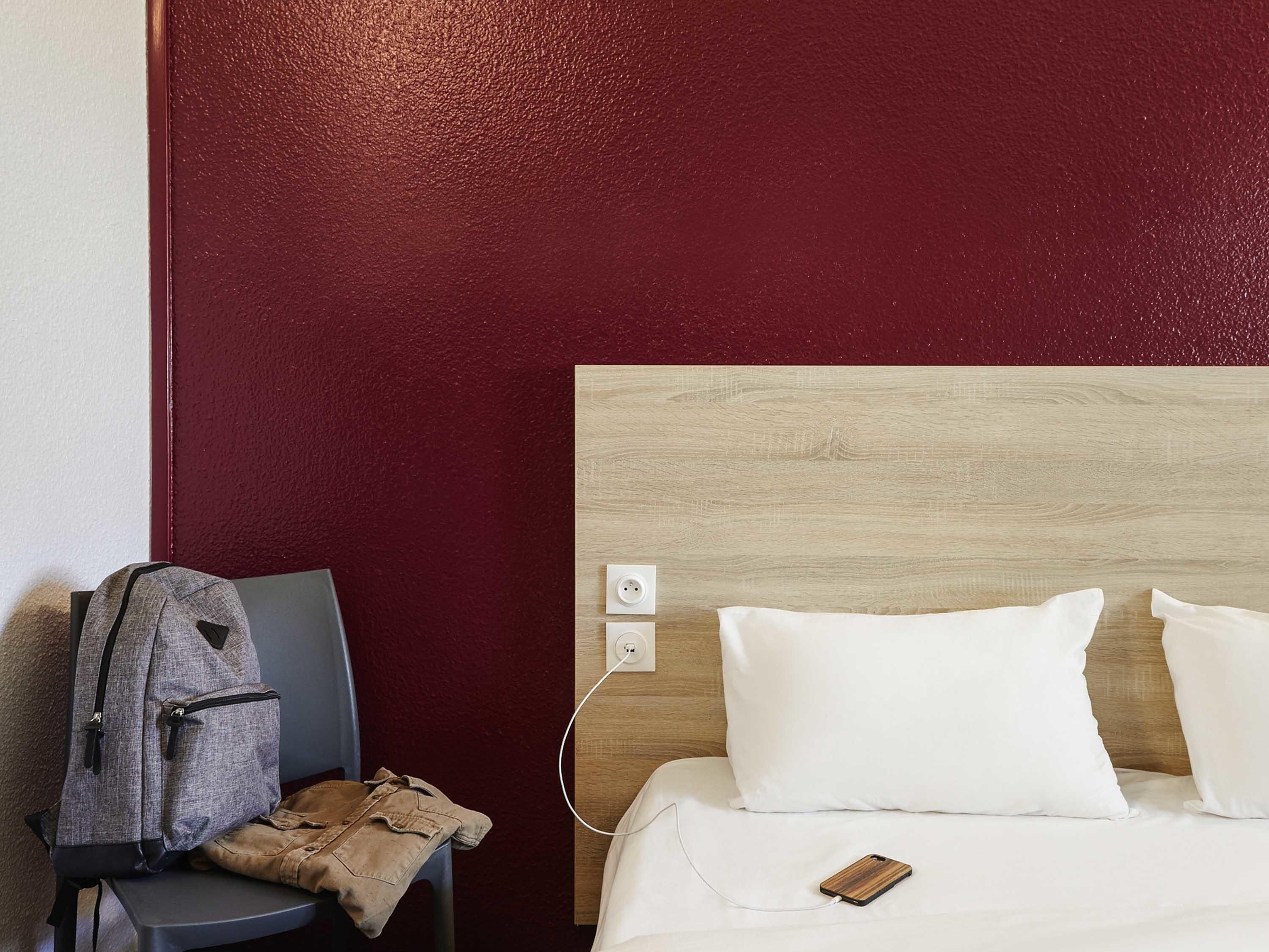 Una habitación de hotel con un cabecero de madera, una mochila gris sobre una silla, un teléfono de madera en la cama y una pared roja.