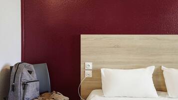 Una habitación de hotel con un cabecero de madera, una mochila gris sobre una silla, un teléfono de madera en la cama y una pared roja.